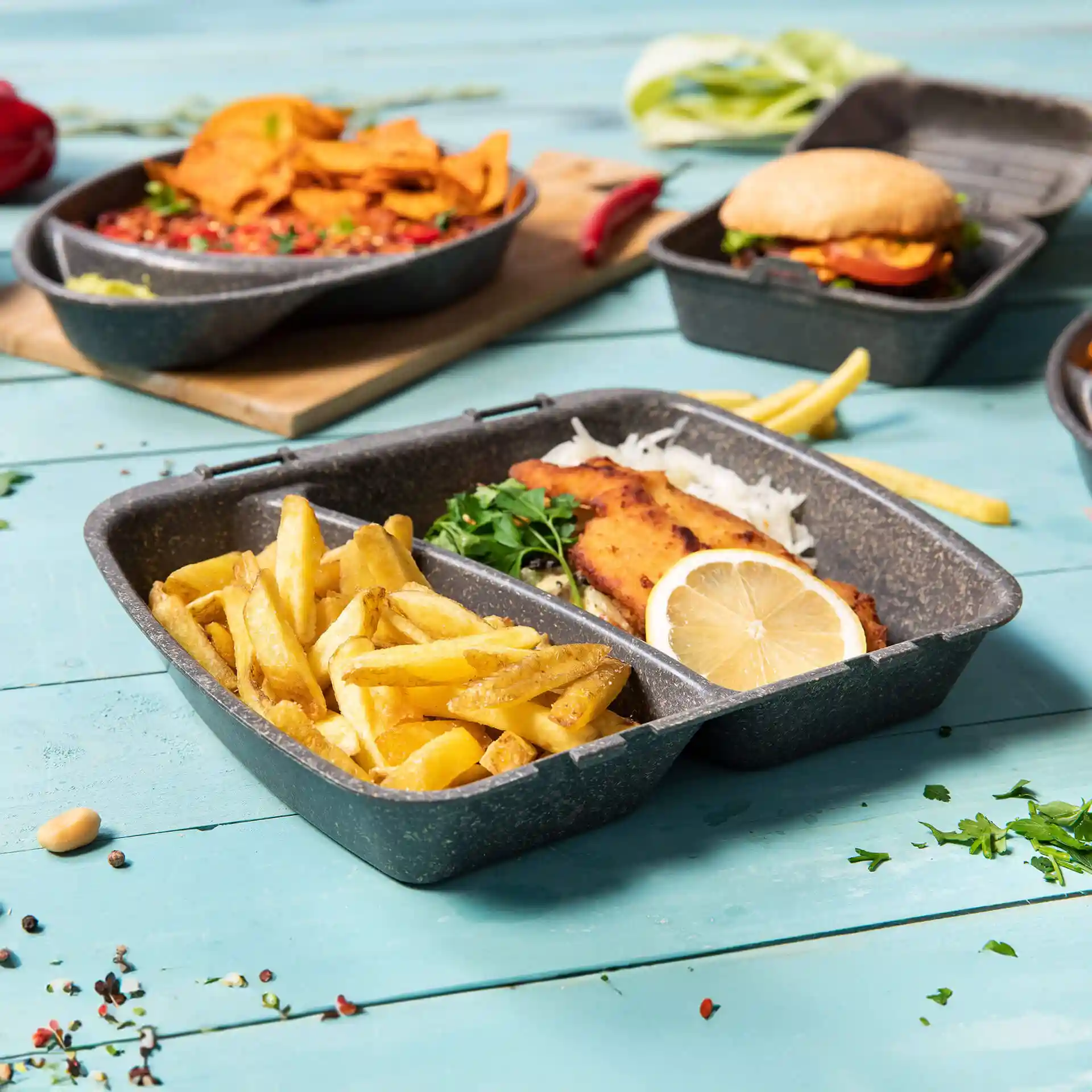 Wiederverwendbarer grauer Essensbehälter mit zwei Fächern, gefüllt mit Pommes und Fisch, Zitronenscheibe und Kräutern.