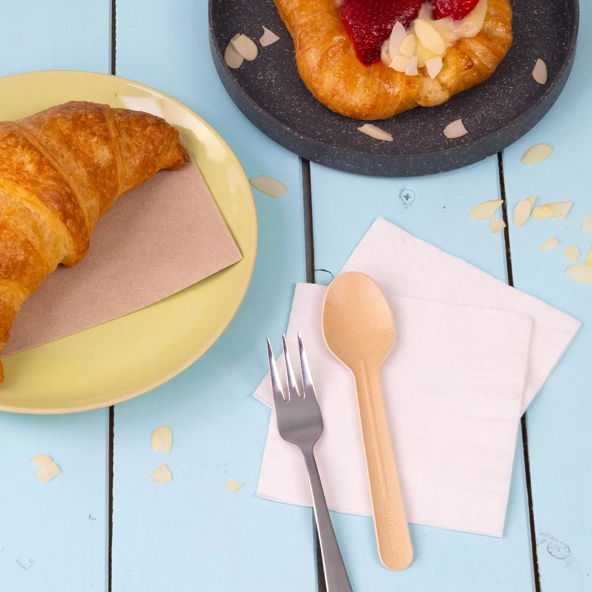 Bild eines Croissants auf einem gelben Teller, einem Gebäck auf einem dunklen Teller, einem Holzlöffel, einer silbernen Gabel und weißen Servietten.