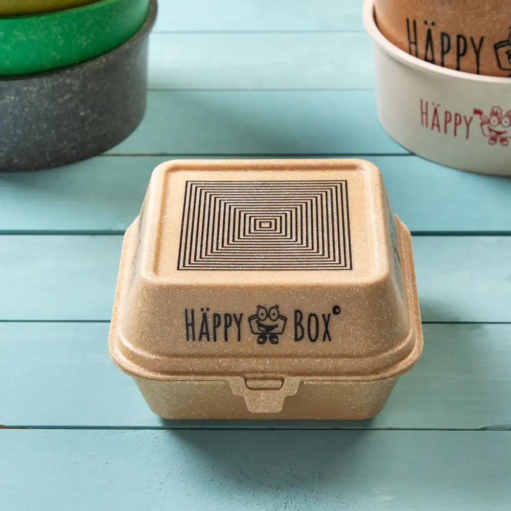 Quadratische Burgerbox in Beige mit geometrischem Design oben und "HÄPPY BOX"-Logo an der Seite, umgeben von bunten Behältern.