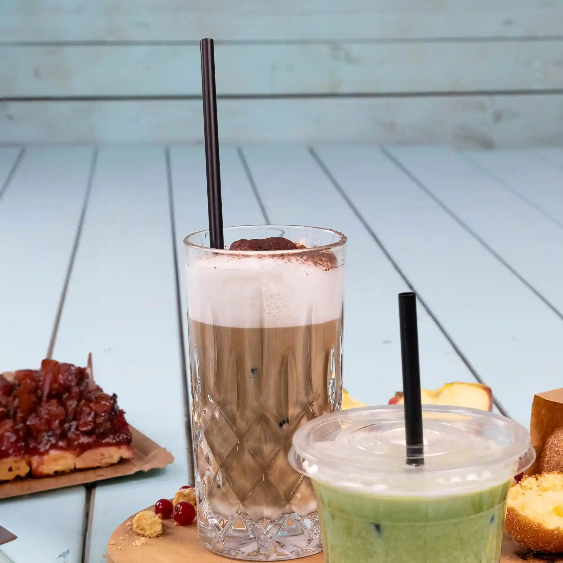 Image of two drinks with black reusable straws, one in a clear glass and another in a plastic cup, on a wooden table.