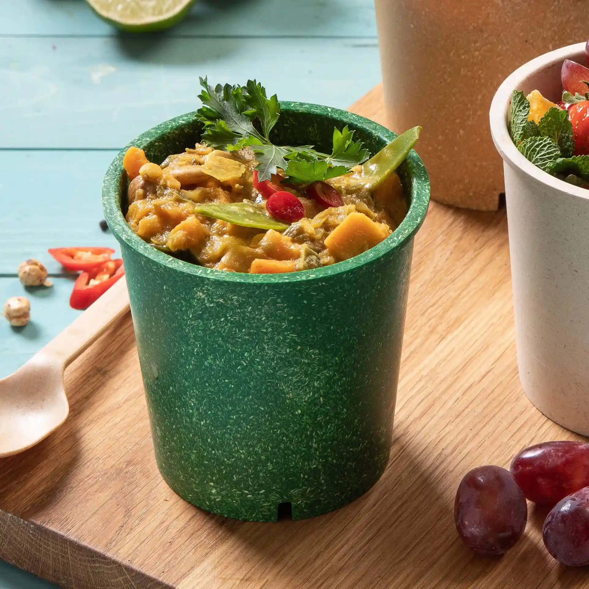 Grüner wiederverwendbarer Becher, gefüllt mit Essen, garniert mit Koriander, rotem Chili und grünen Bohnen.