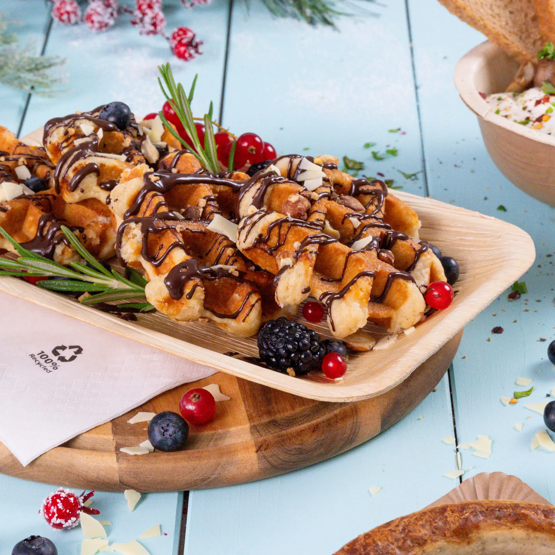 Plate of waffles drizzled with chocolate, garnished with berries and rosemary, on a palm leaf disposable plate.