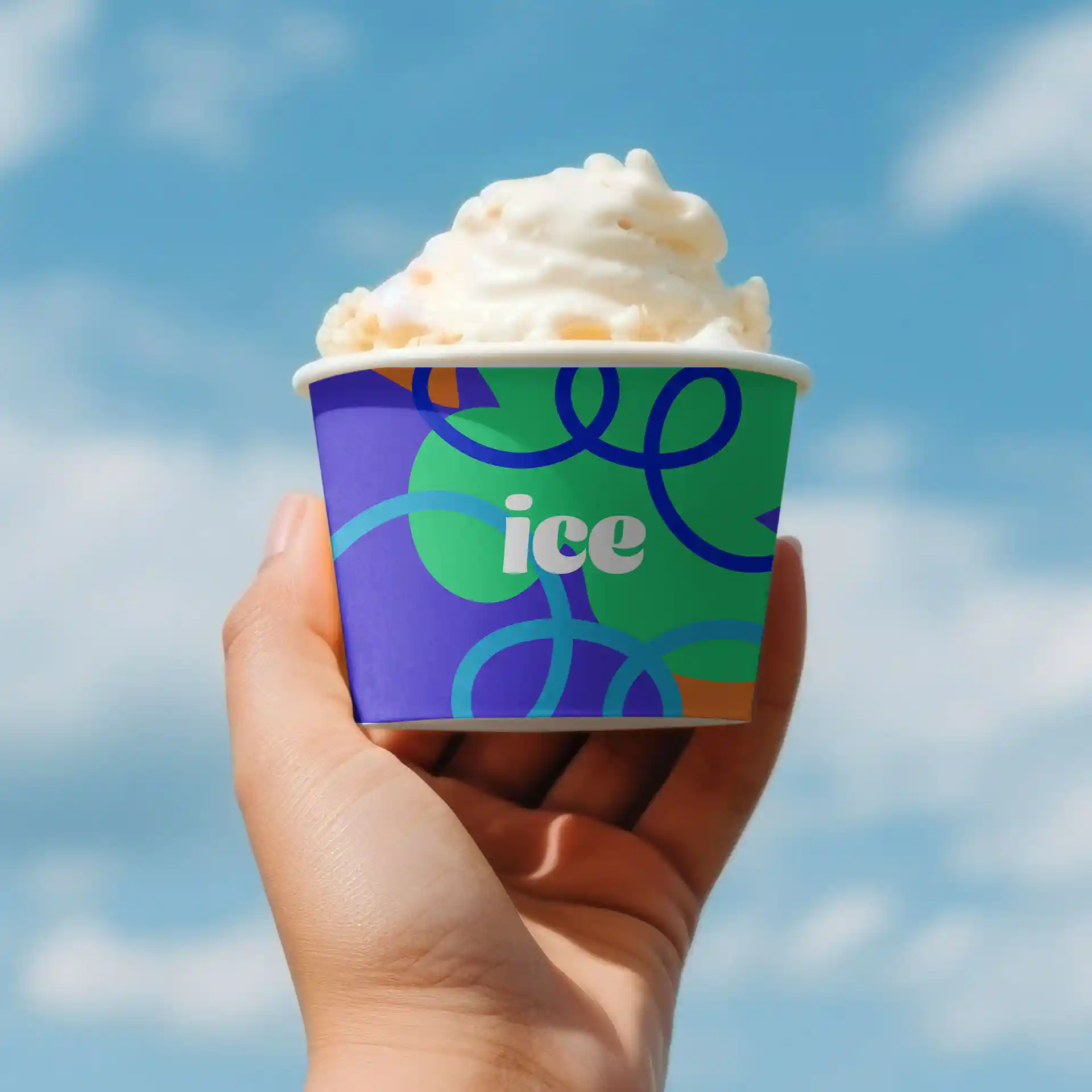 Hand holding a round paperboard ice cream cup with a colorful design, filled with soft serve ice cream against a blue sky.