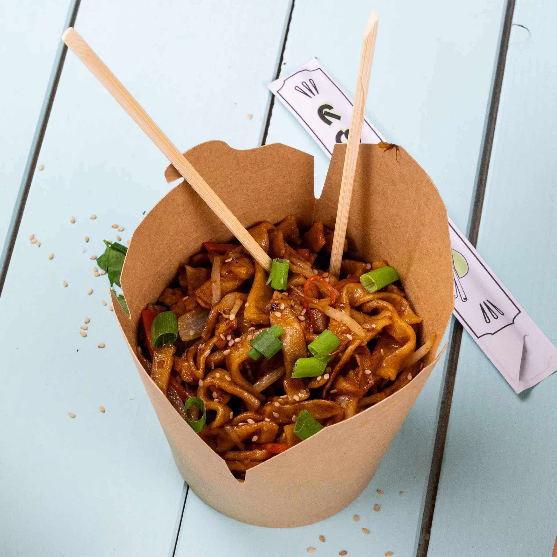 Noodle box filled with fried noodles topped with green onions and sesame seeds, held by chopsticks on a blue surface.