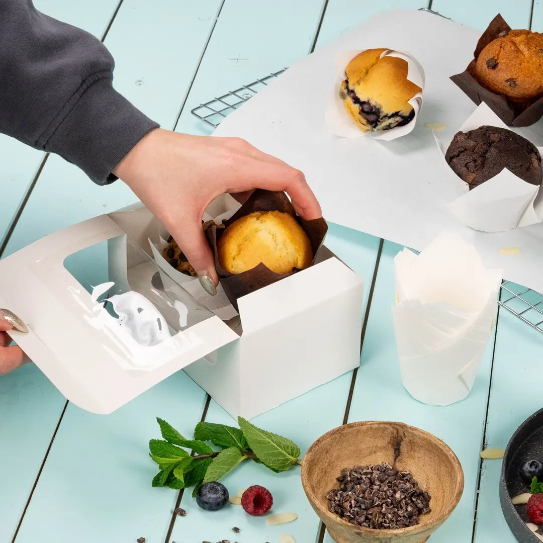 Hand placing a muffin into a white cupcake box with several baked goods and ingredients on a blue surface.