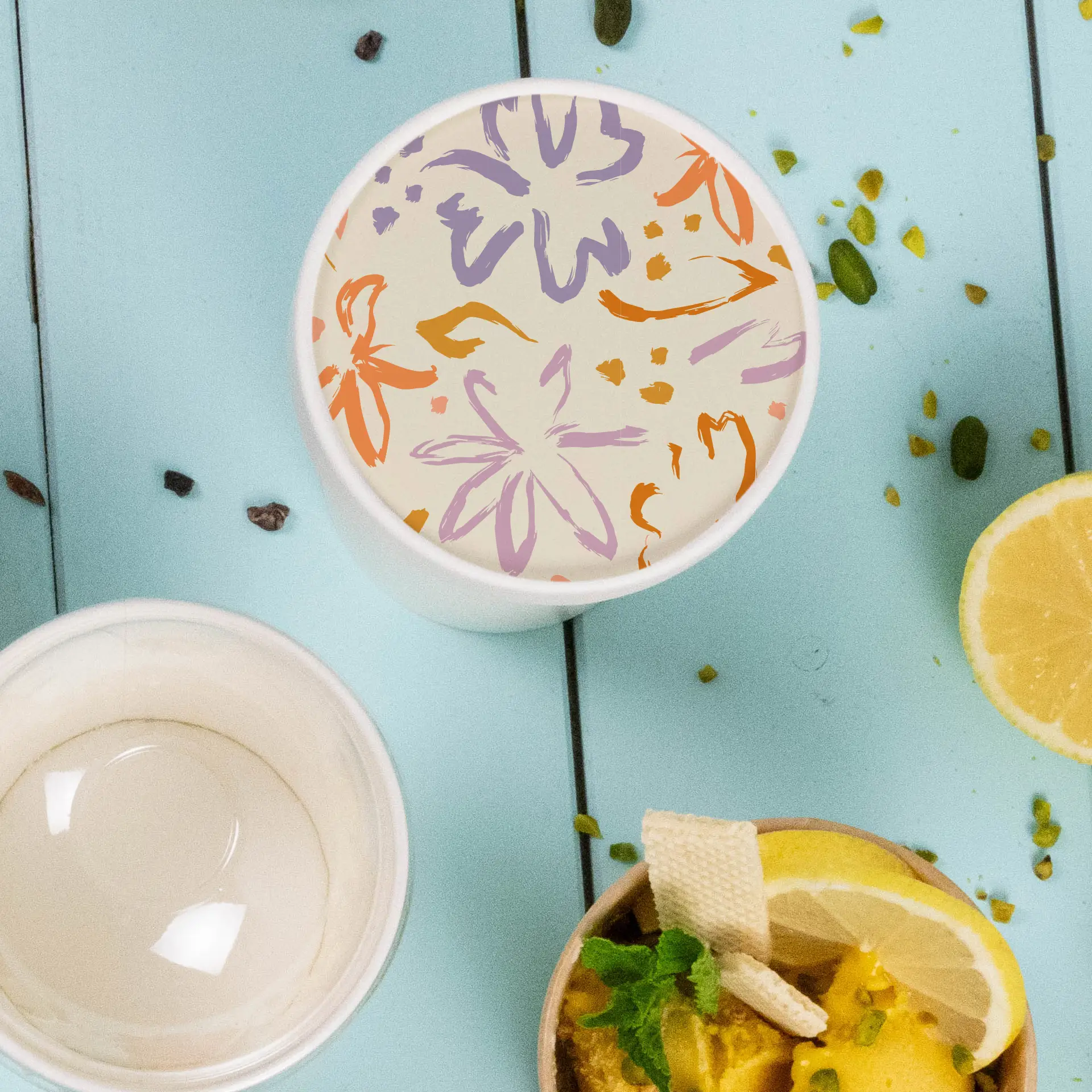 Individuell bedruckter Deckel für Becher mit floralen Motiven, auf einer blauen Fläche mit Lebensmitteln daneben.