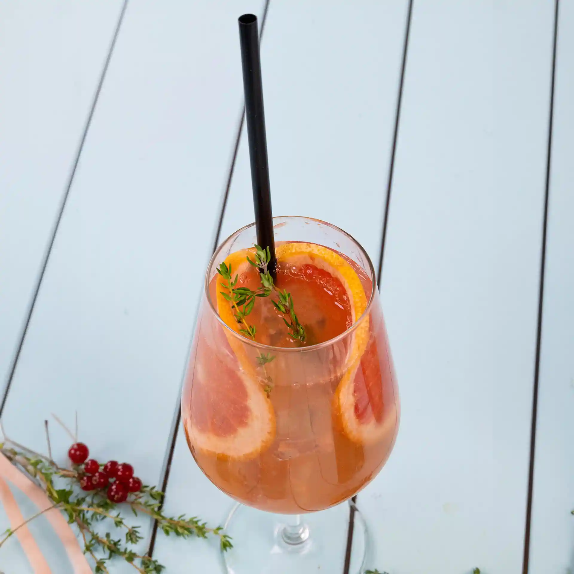 Image of a clear glass filled with a drink, garnished with orange slices and thyme, with a black straw.