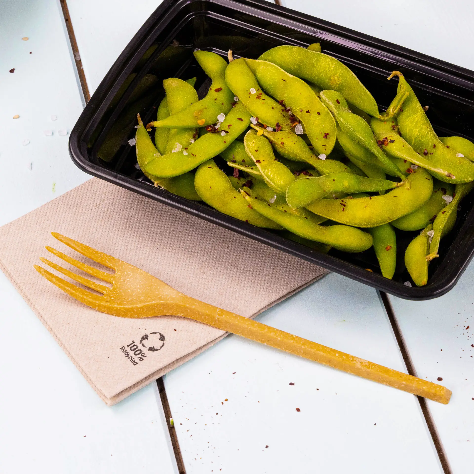 A black container filled with green edamame garnished with spices, accompanied by a biodegradable fork on a recycled paper napkin.