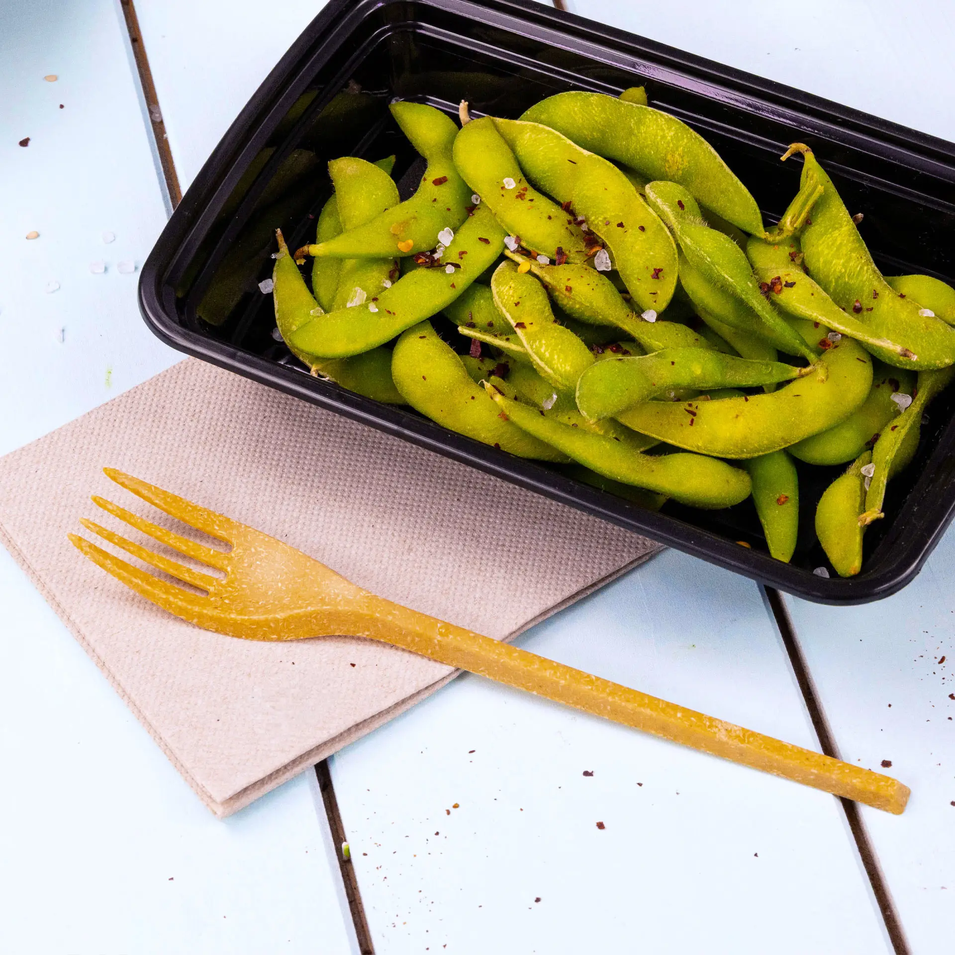 Image of a black container filled with green edamame beans, a yellow fork on a beige napkin beside it.