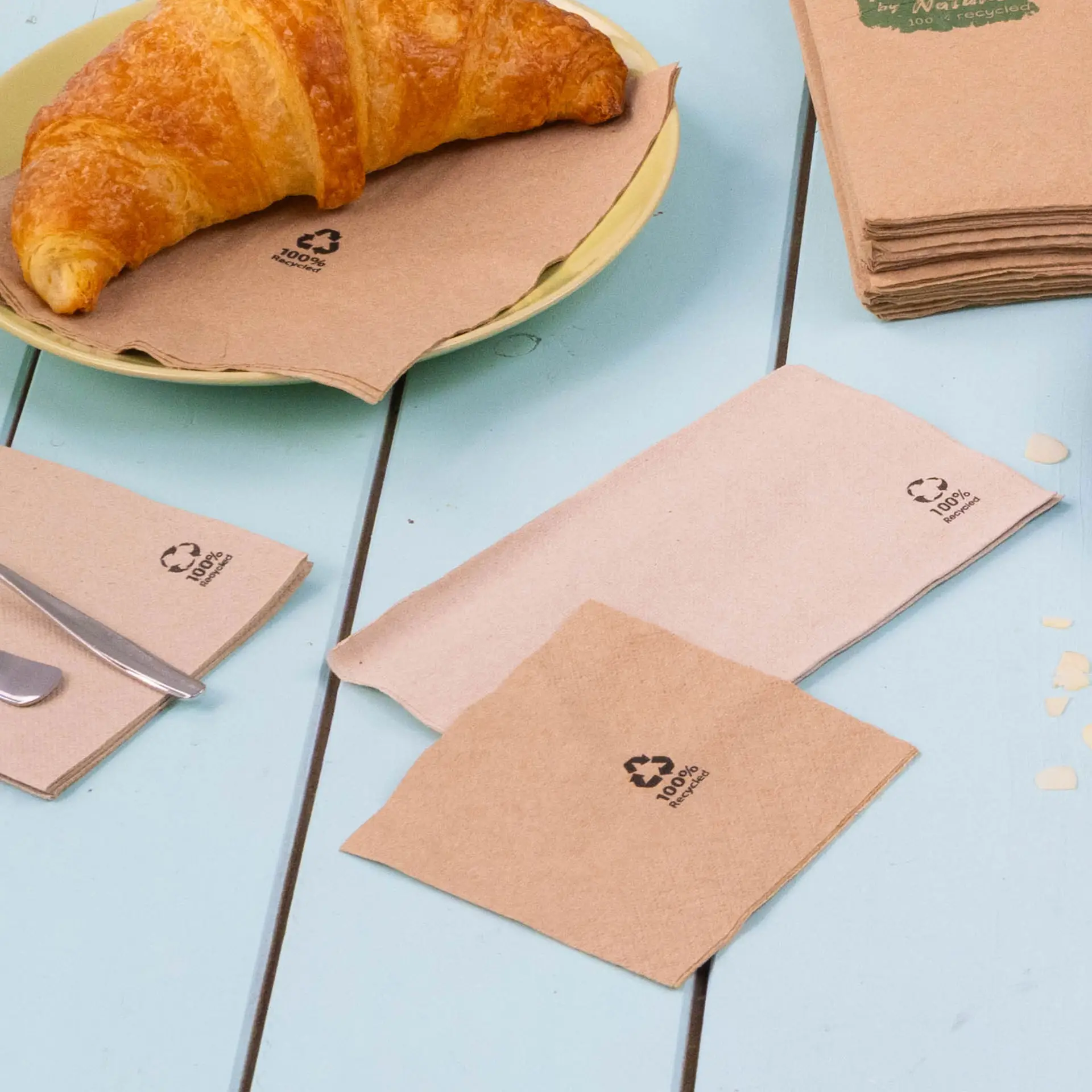 Image of brown, biodegradable cocktail napkins on a blue table, with a croissant on a plate, showcasing recycling symbols.