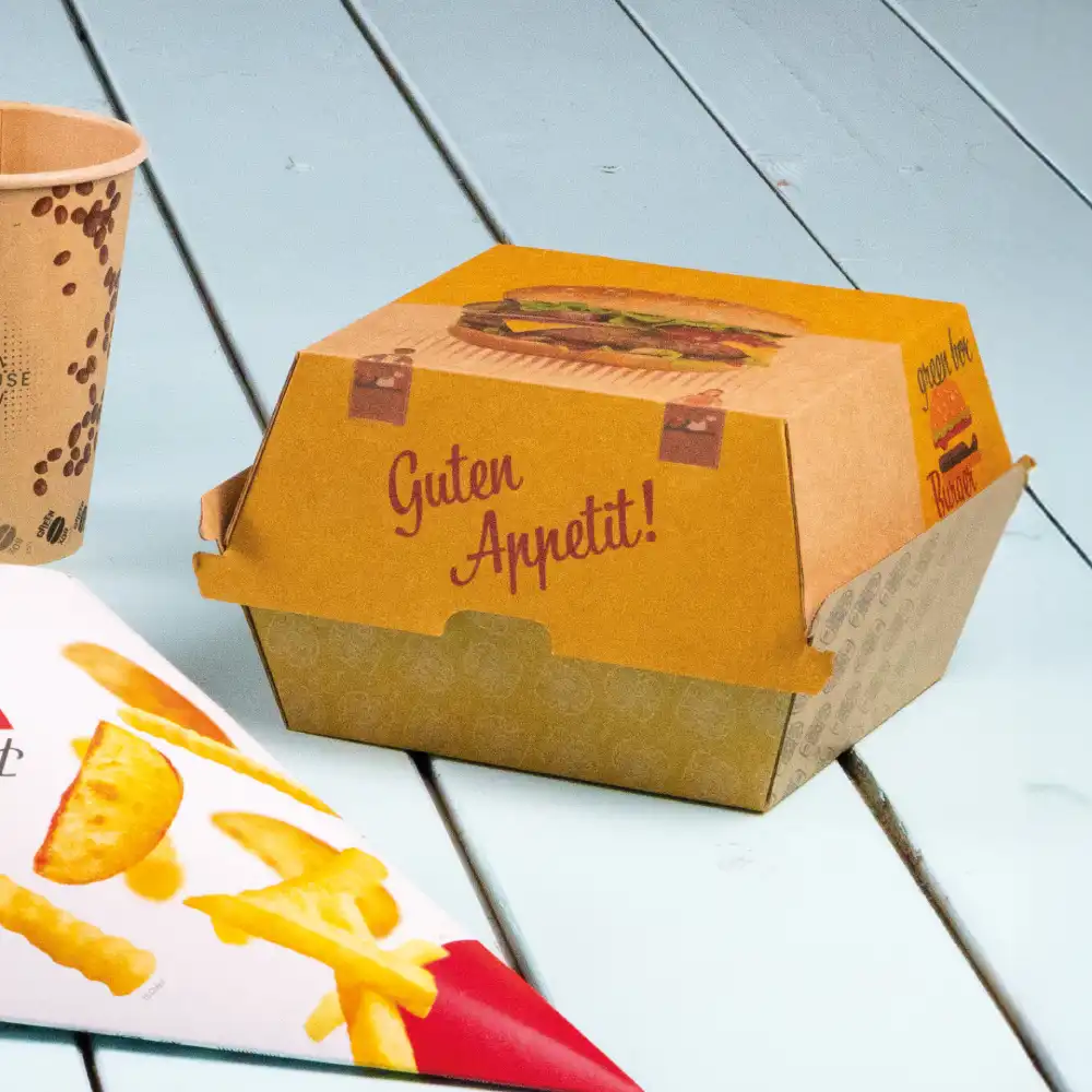 Fastfood-Box mit Burger-Illustration und "Guten Appetit!" Text; Pappbecher und Pommes-Verpackung daneben auf einer Holzoberfläche.