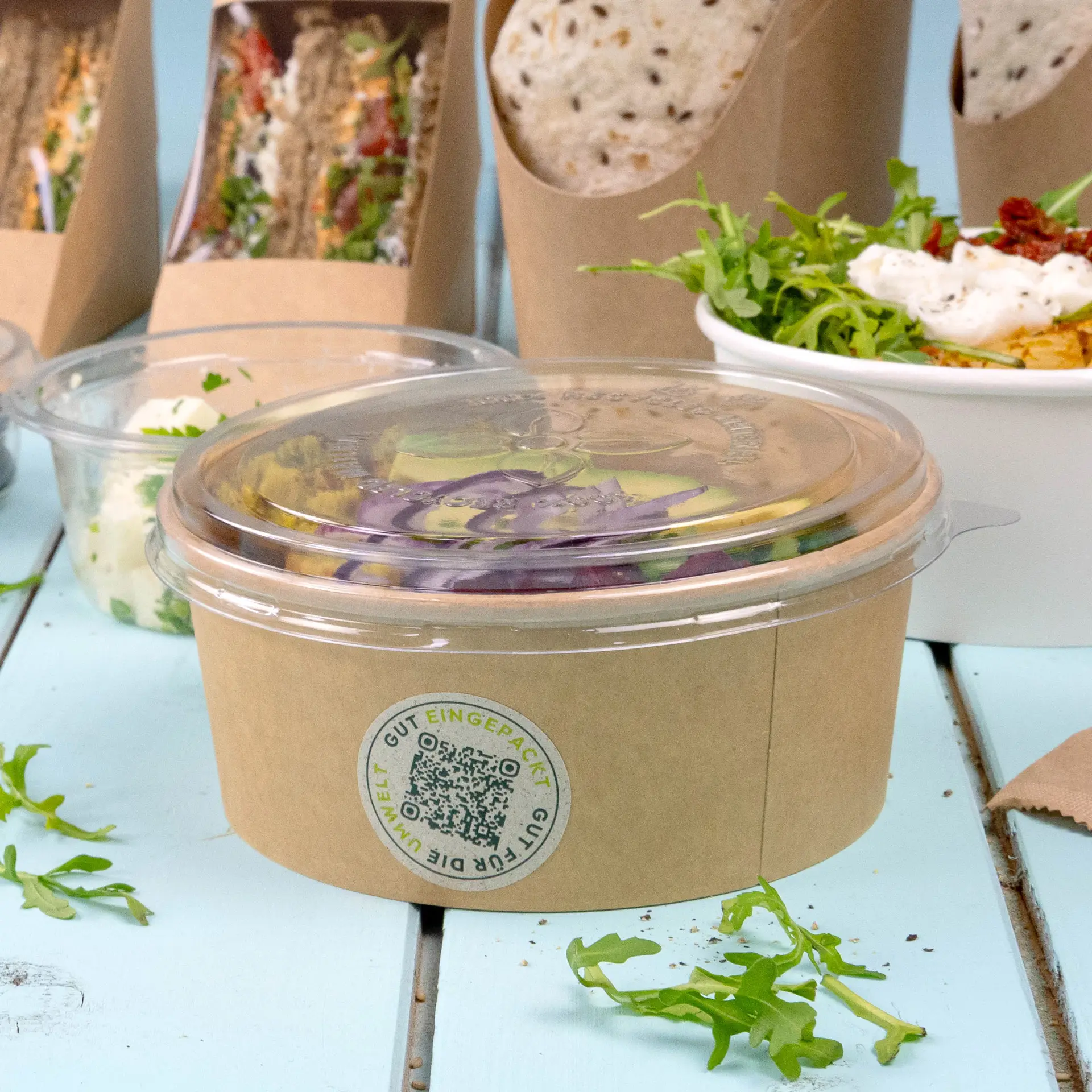 One clear rPET lid for a deli container; surrounded by packaged food items and fresh greens on a light blue surface.