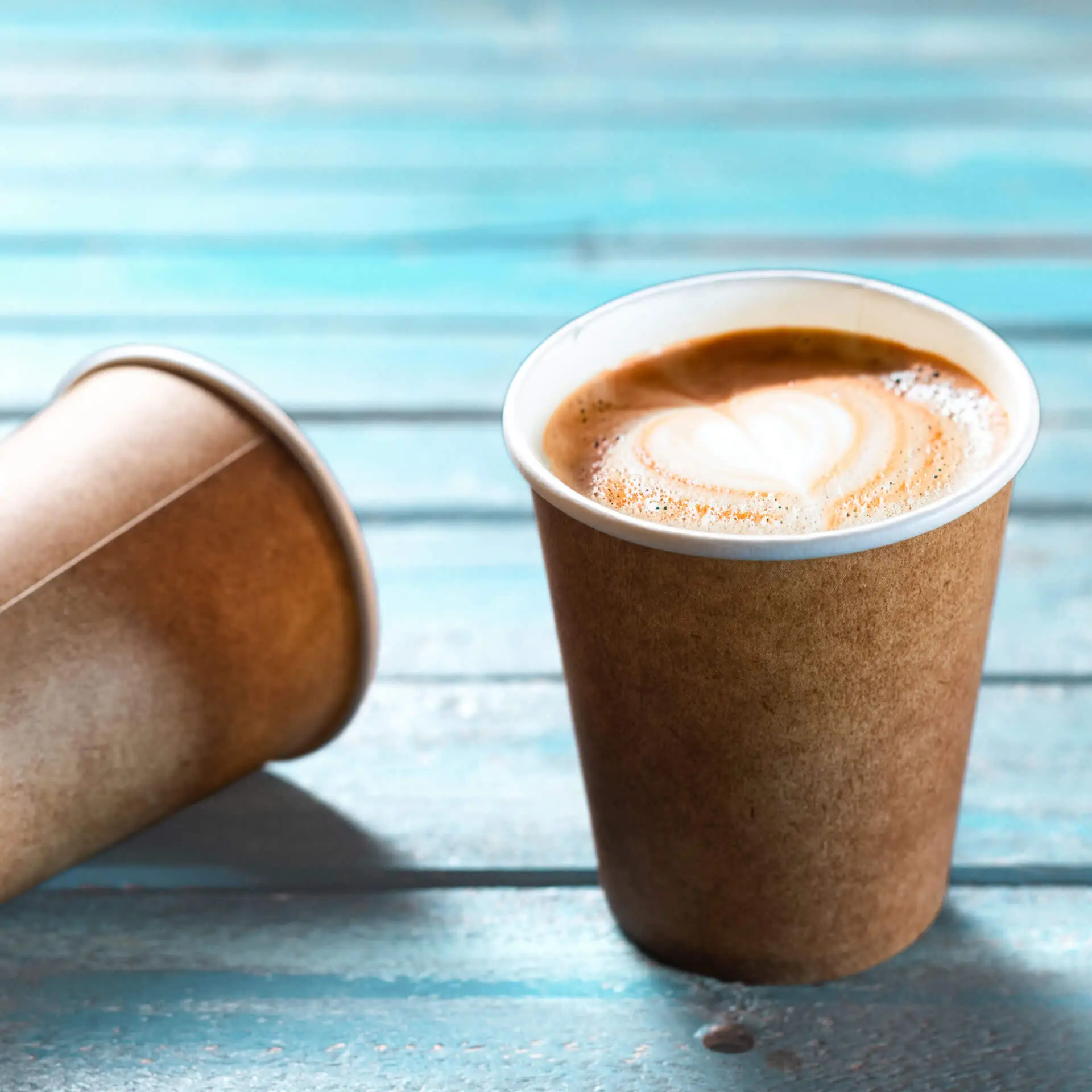 Brauner Becher mit weißem Innenfutter, gefüllt mit Kaffee und Herz-Latte-Art, auf einer blauen Holzoberfläche.