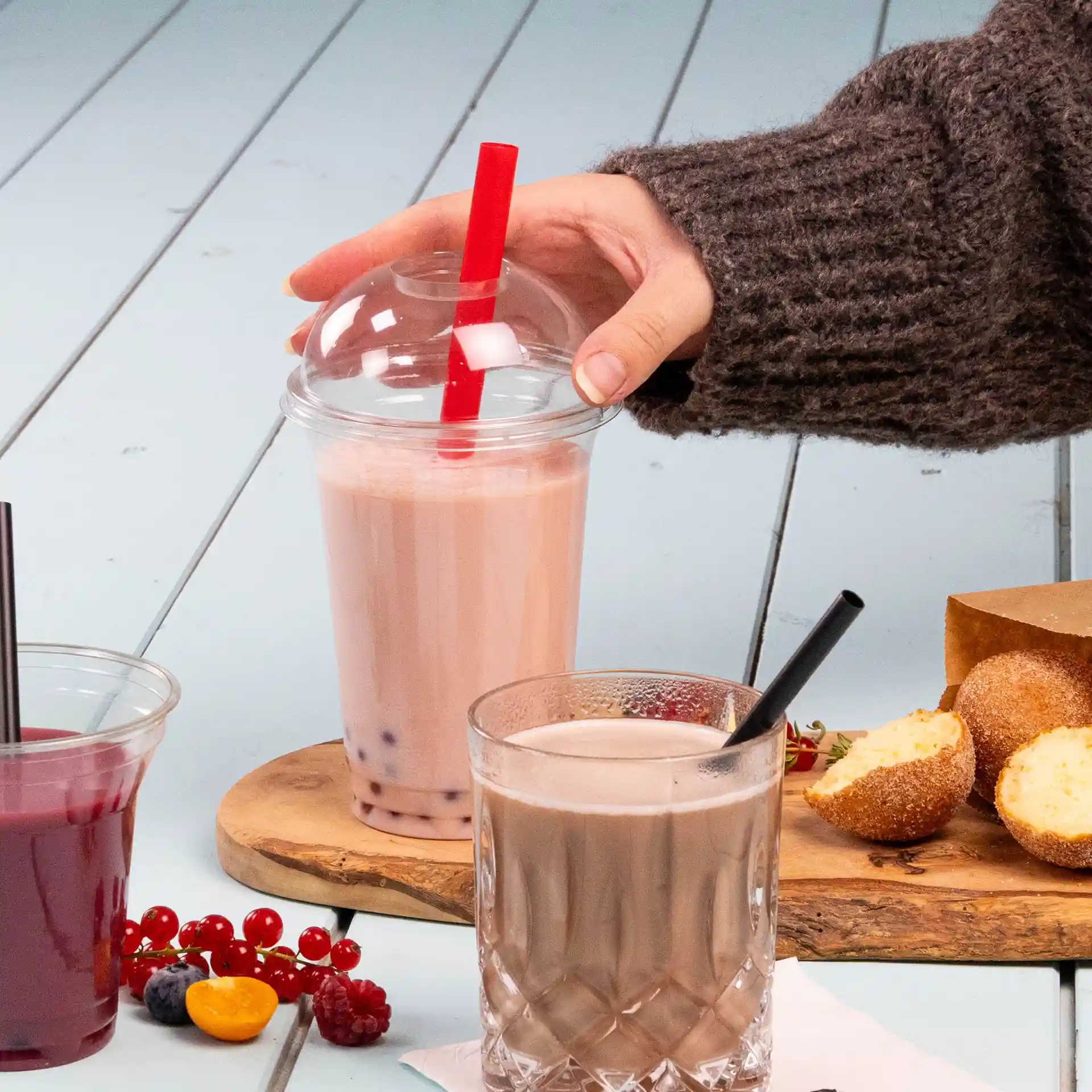 Image of a hand holding a bubble tea cup with a red straw, next to a glass of chocolate drink and snacks on a wooden board.