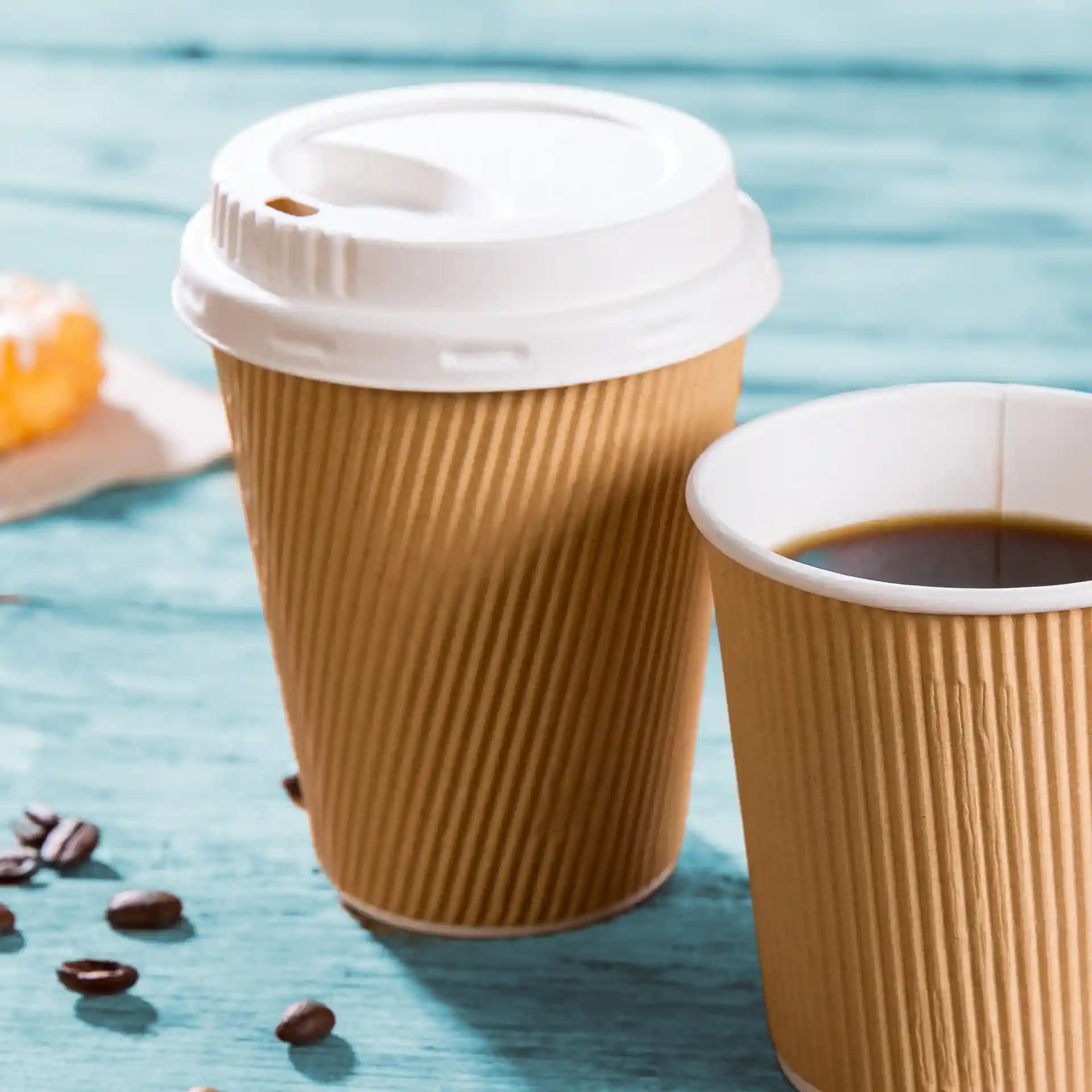 Two brown ripple coffee cups, one with a white lid, placed on a blue wooden surface with coffee beans nearby.
