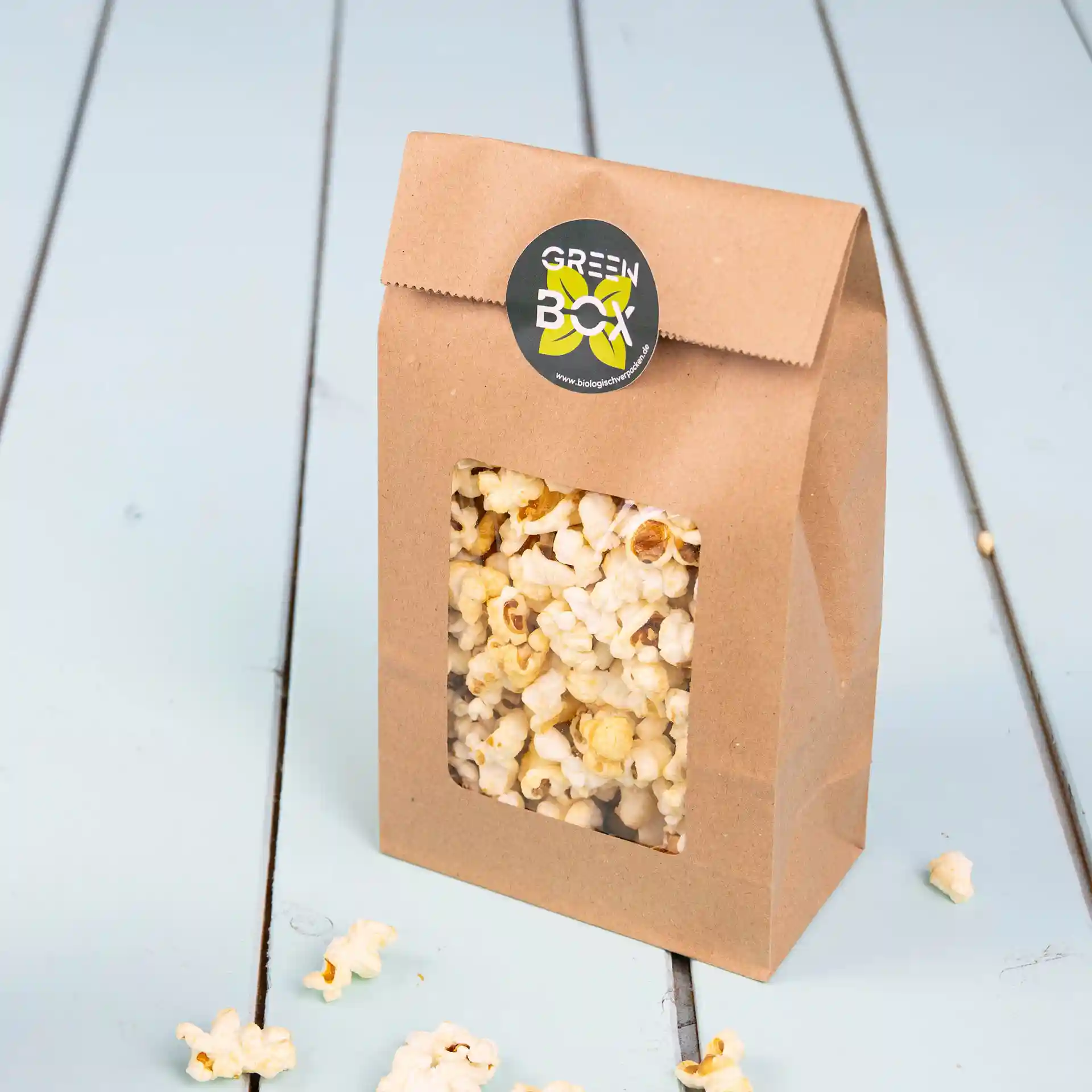 Braune Kraftpapiertüte mit transparentem Fenster, gefüllt mit Popcorn, auf einer hellblauen Holzoberfläche.