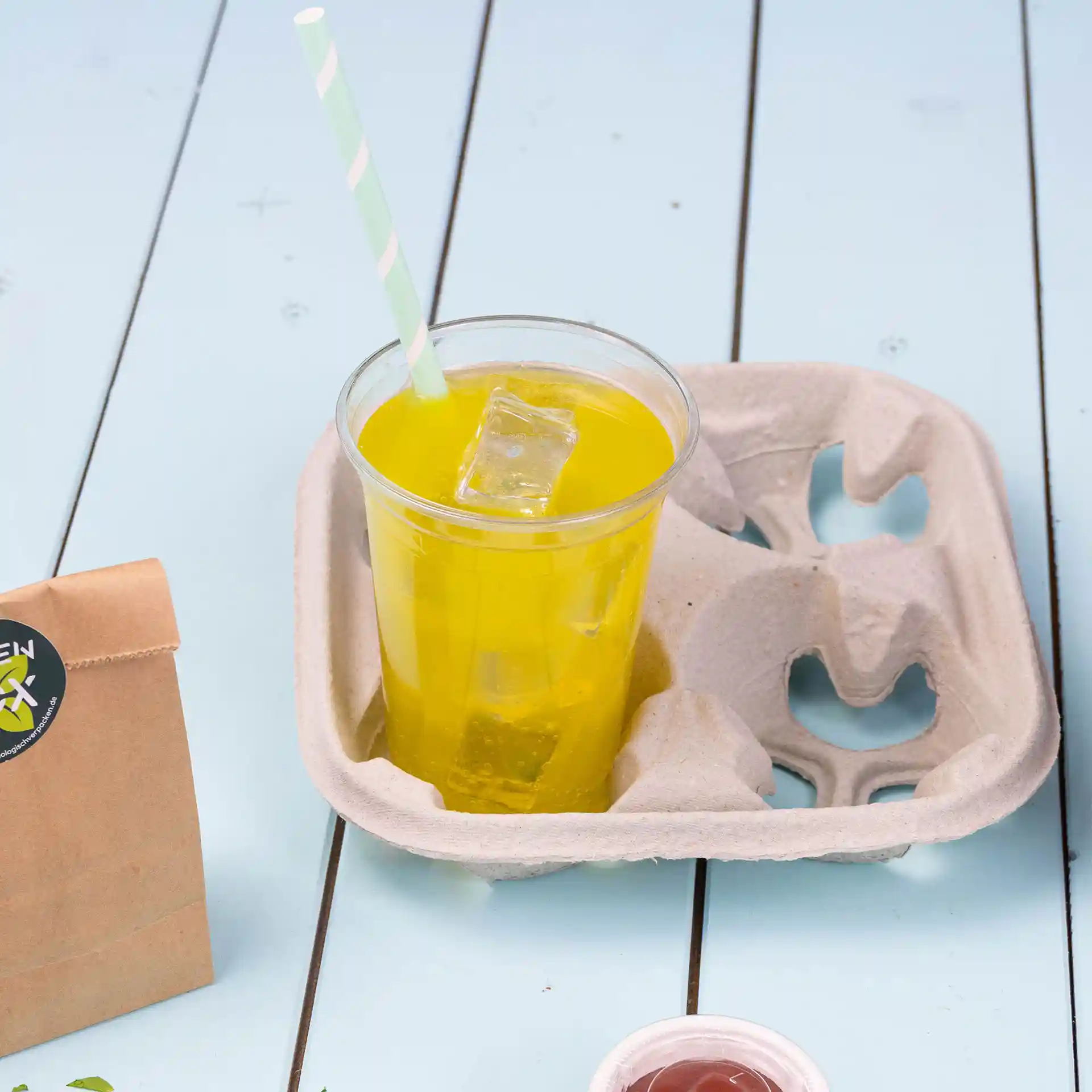 Cup holder made of unbleached paperboard with a yellow drink and a green striped straw, ice cubes visible inside.