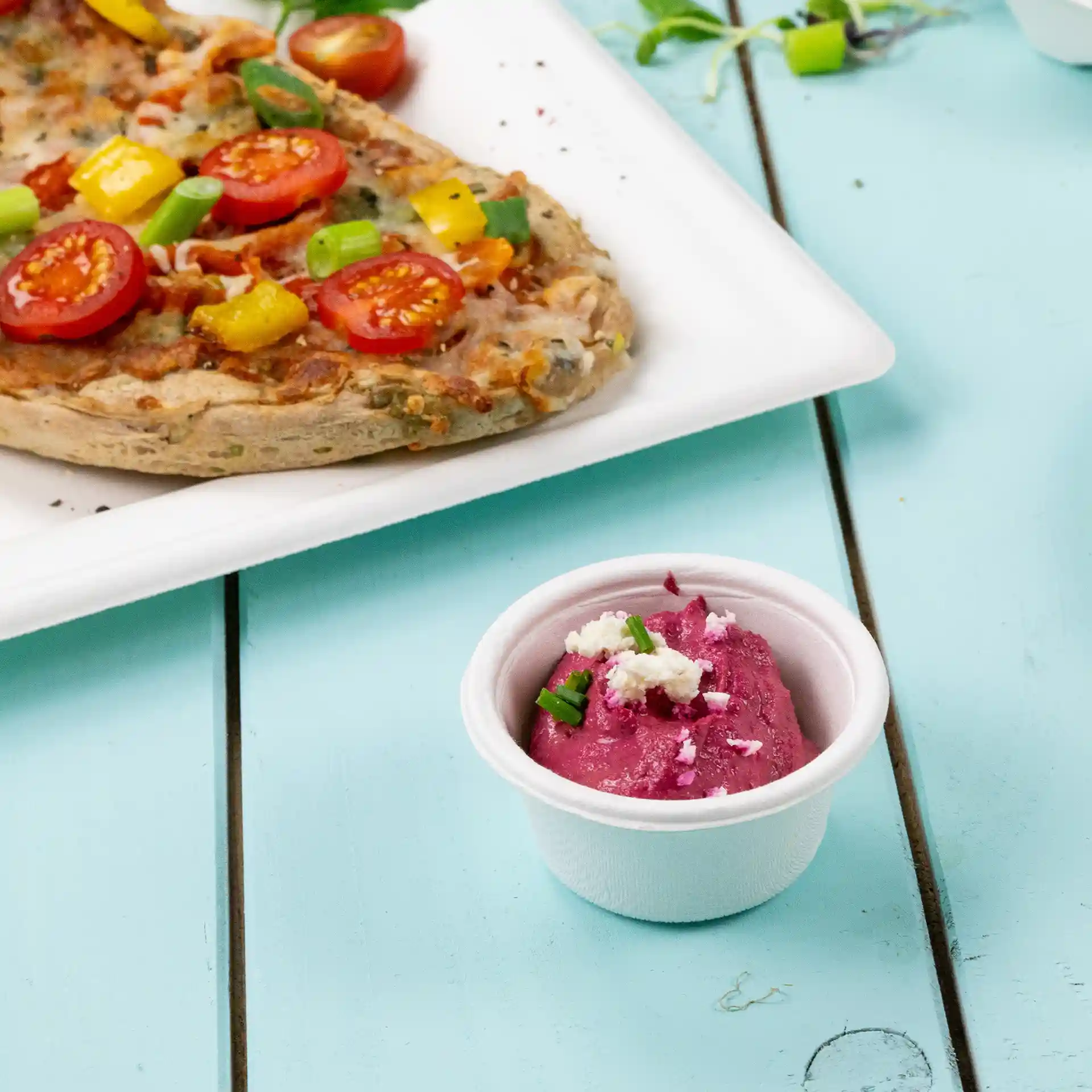 White bagasse dip bowl with purple dip and green garnish, placed next to a pizza on a white plate.