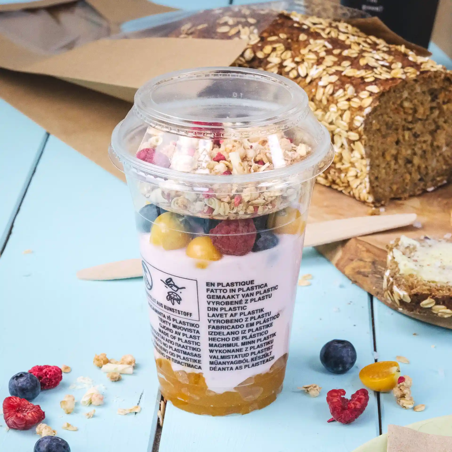 Transparent 12 oz PLA smoothie cup filled with yogurt, granola, and mixed fruits, placed on a blue surface with bread in the background.