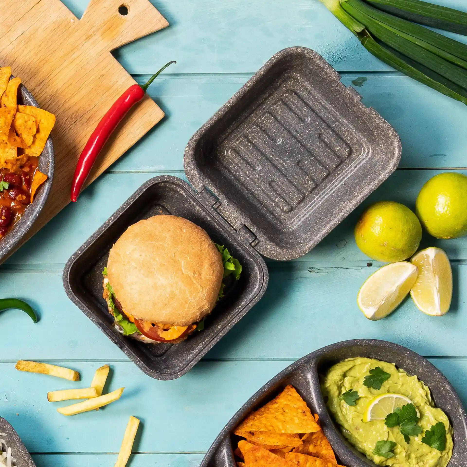 Wiederverwendbare graue Burgerbox mit Deckel, enthält einen Burger, umgeben von Limette, Nachos und einer Chilischote.