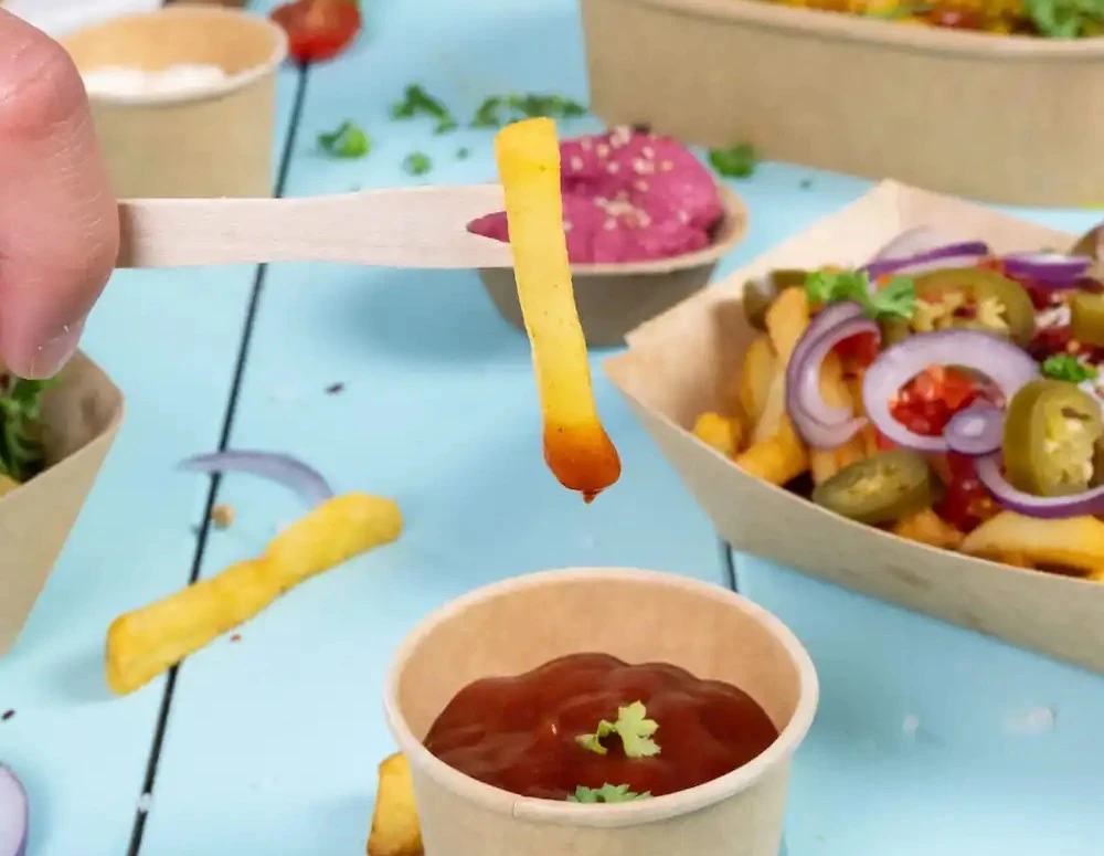 A hand holds a wooden stick with a French fry above a cup of ketchup placed on a blue surface.