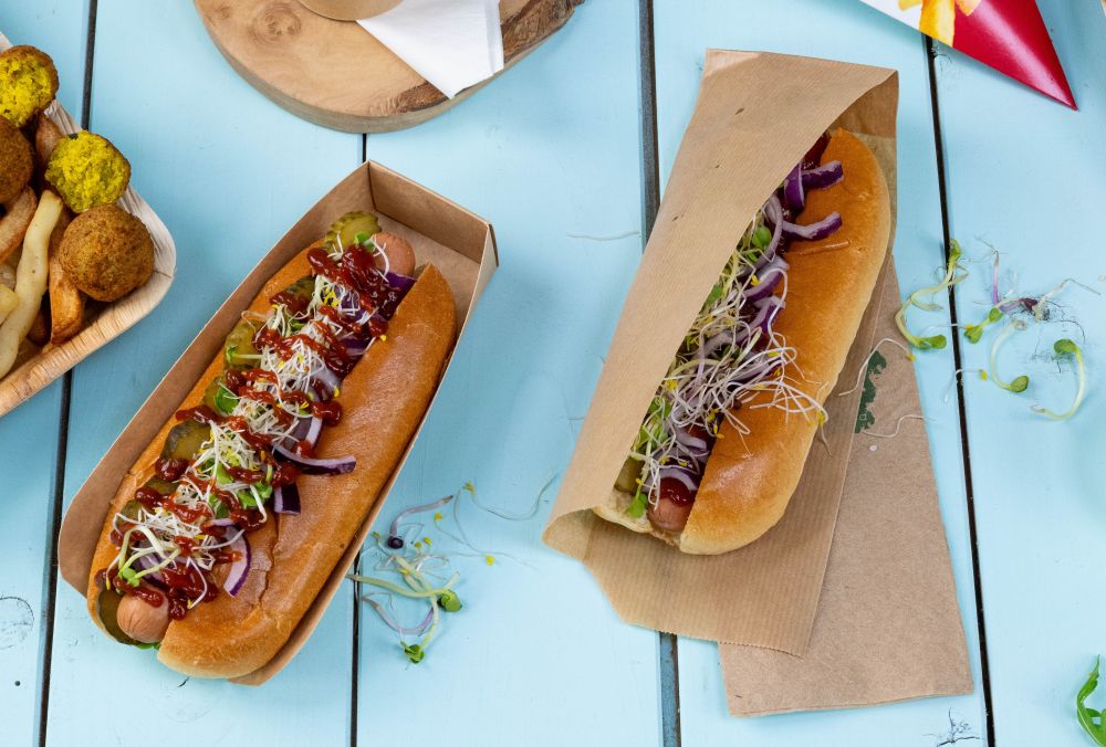Zwei Hotdogs mit verschiedenen Toppings, darunter Gurken, Zwiebeln und Sprossen, in Papierbehältern auf einem blauen Tisch.