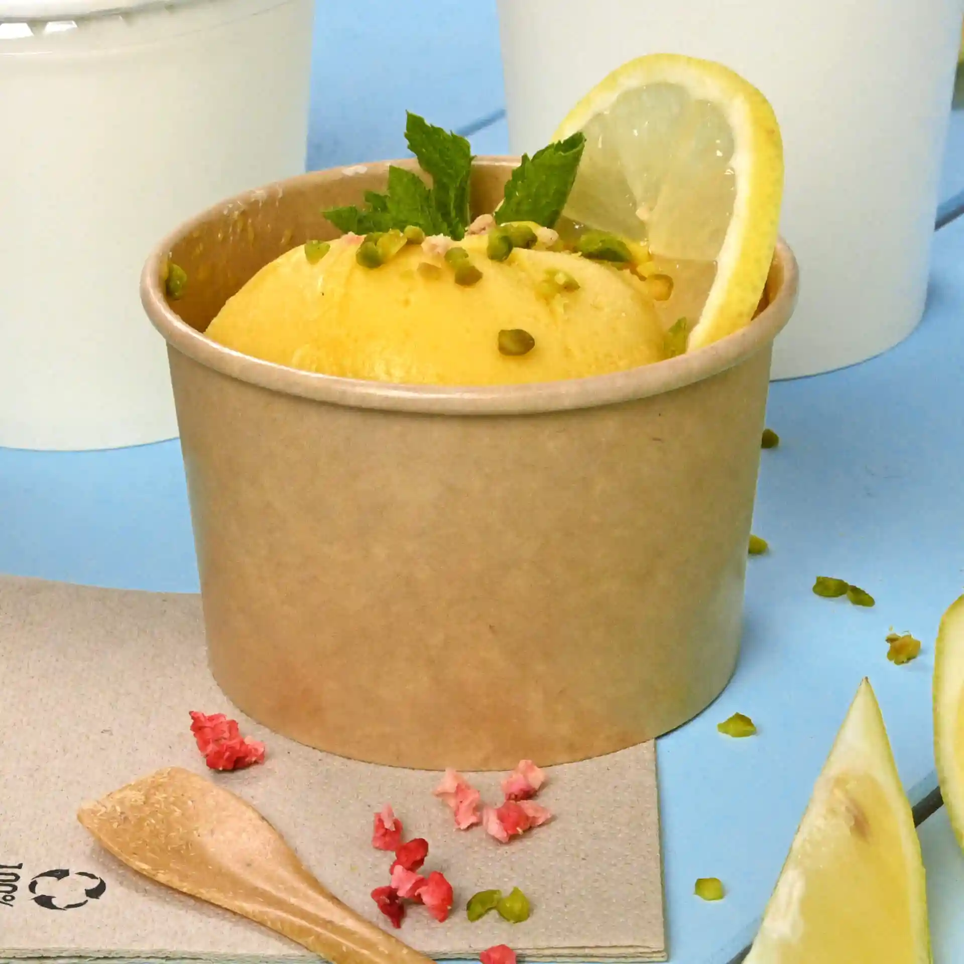 Brauner Pappbecher mit gelbem Eis, garniert mit einer Zitronenscheibe, Minze und Pistazien, auf blauer Oberfläche.