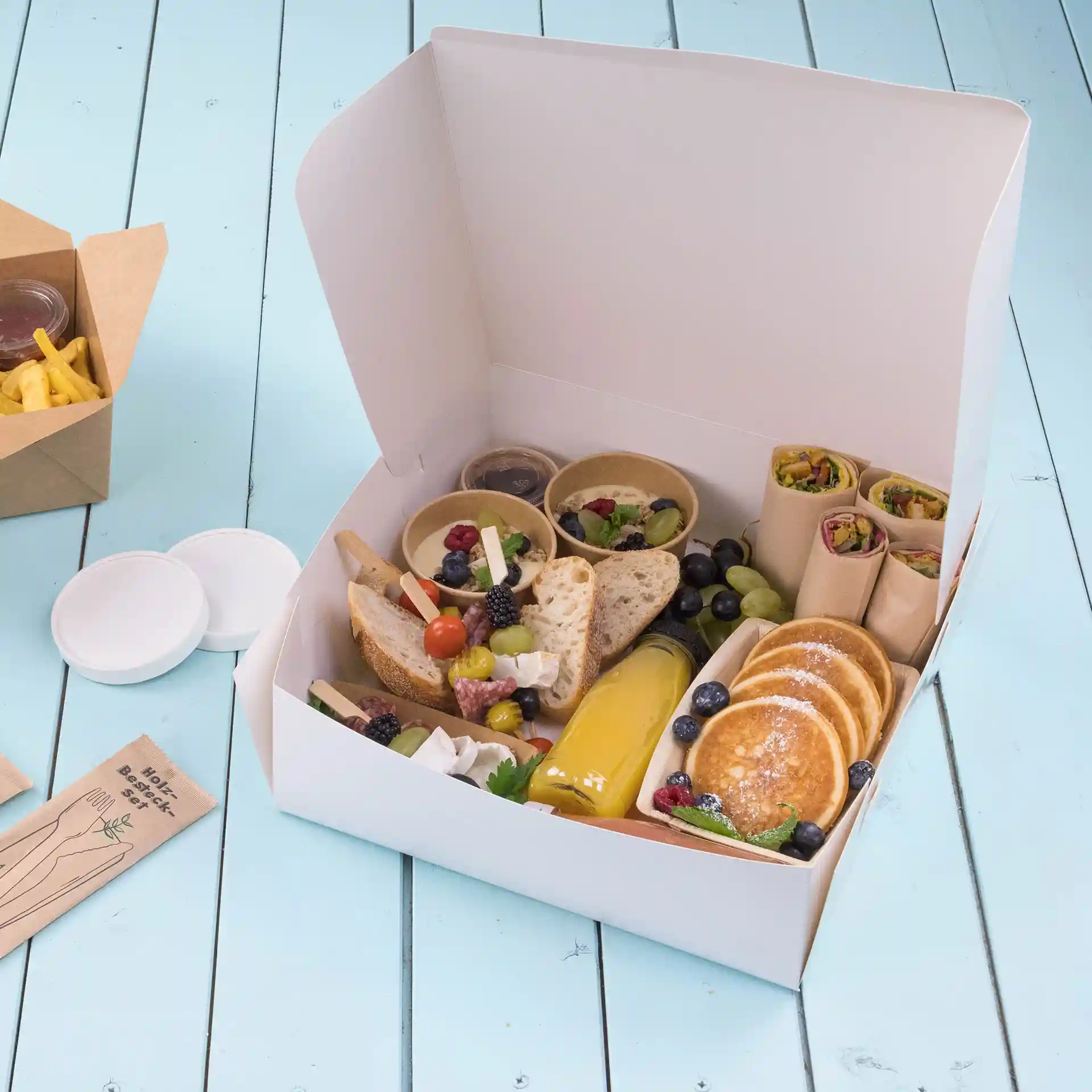 Weiße Kuchenbox gefüllt mit verschiedenen Lebensmitteln wie Brot, Obst, Pfannkuchen und Saucen auf einer blauen Holzoberfläche.
