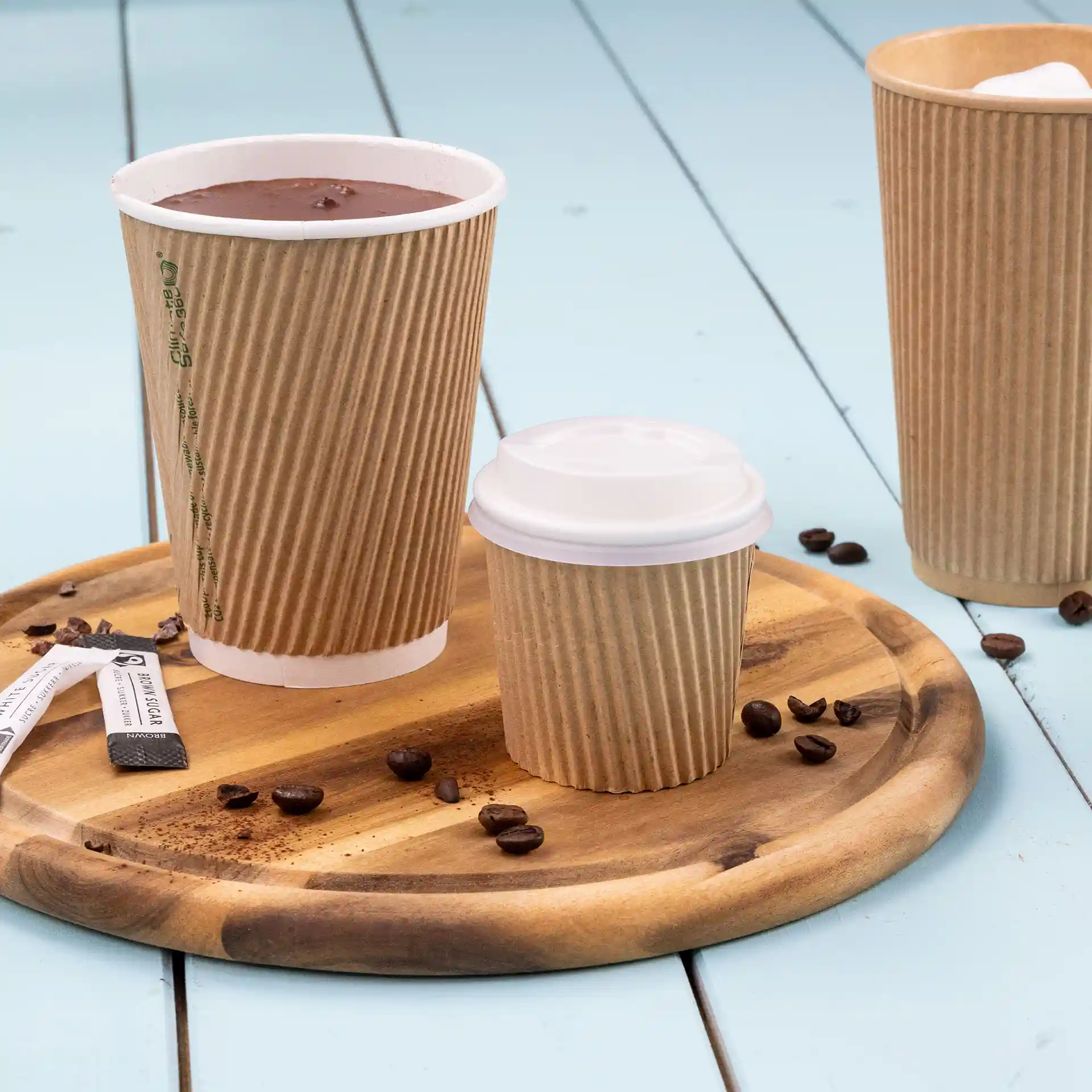 Image of a wooden tray with a large brown ribbed coffee cup, a smaller brown ribbed cup with a white lid, and coffee beans scattered around.