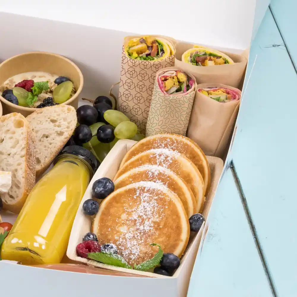 A variety of food items including wraps, pancakes, fruit, and a juice bottle in a takeout box on a blue surface.