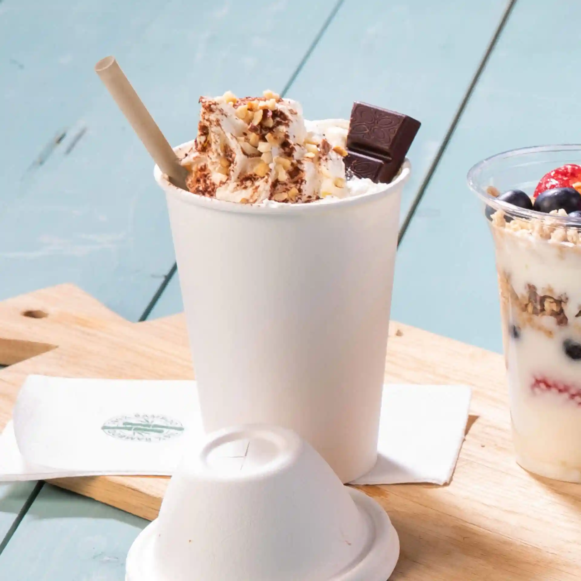 Cup filled with whipped cream, nuts, and chocolate, with a brown paper straw, on a wooden surface.