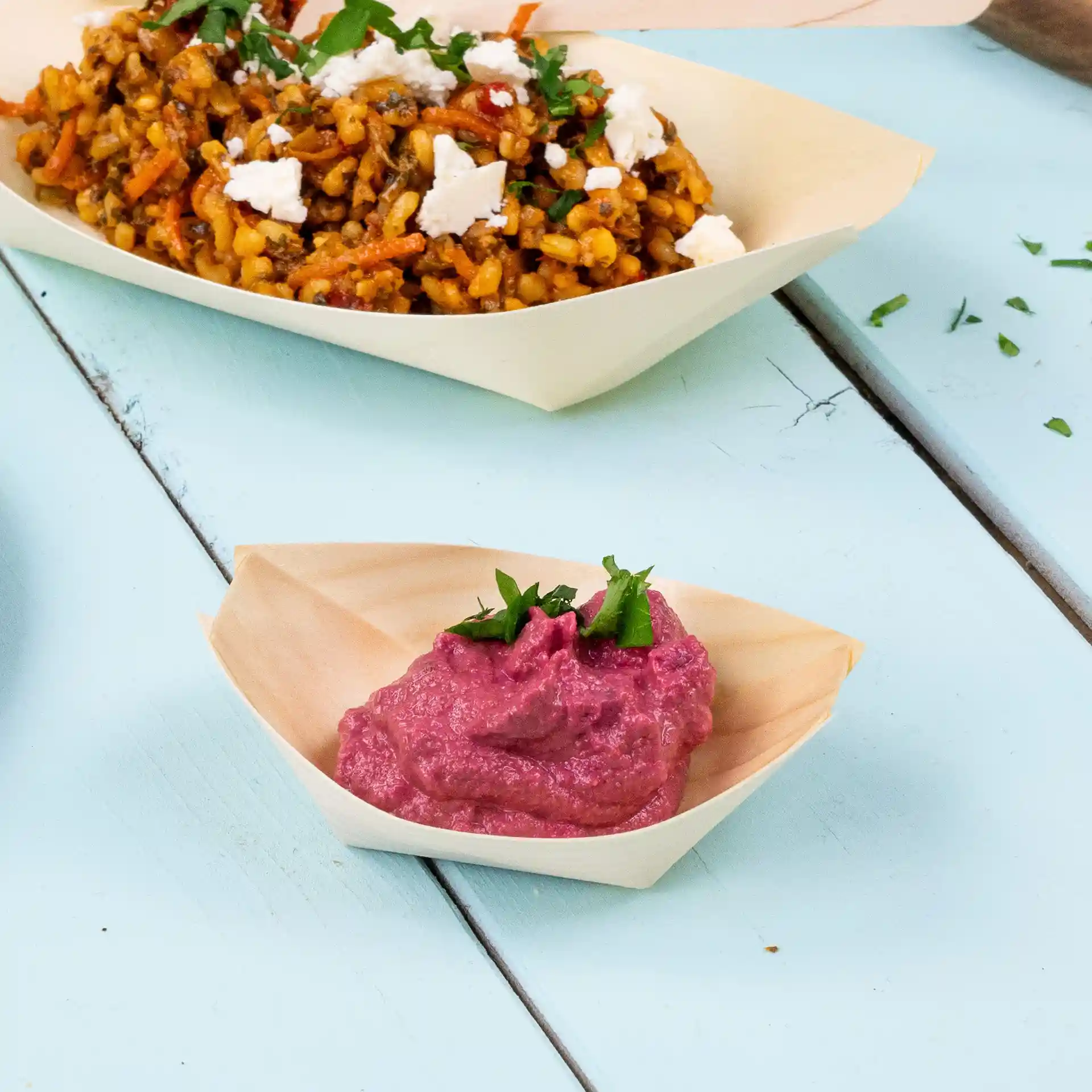 Wooden boat-shaped dish with pink hummus topped with chopped herbs, placed on a light blue surface.