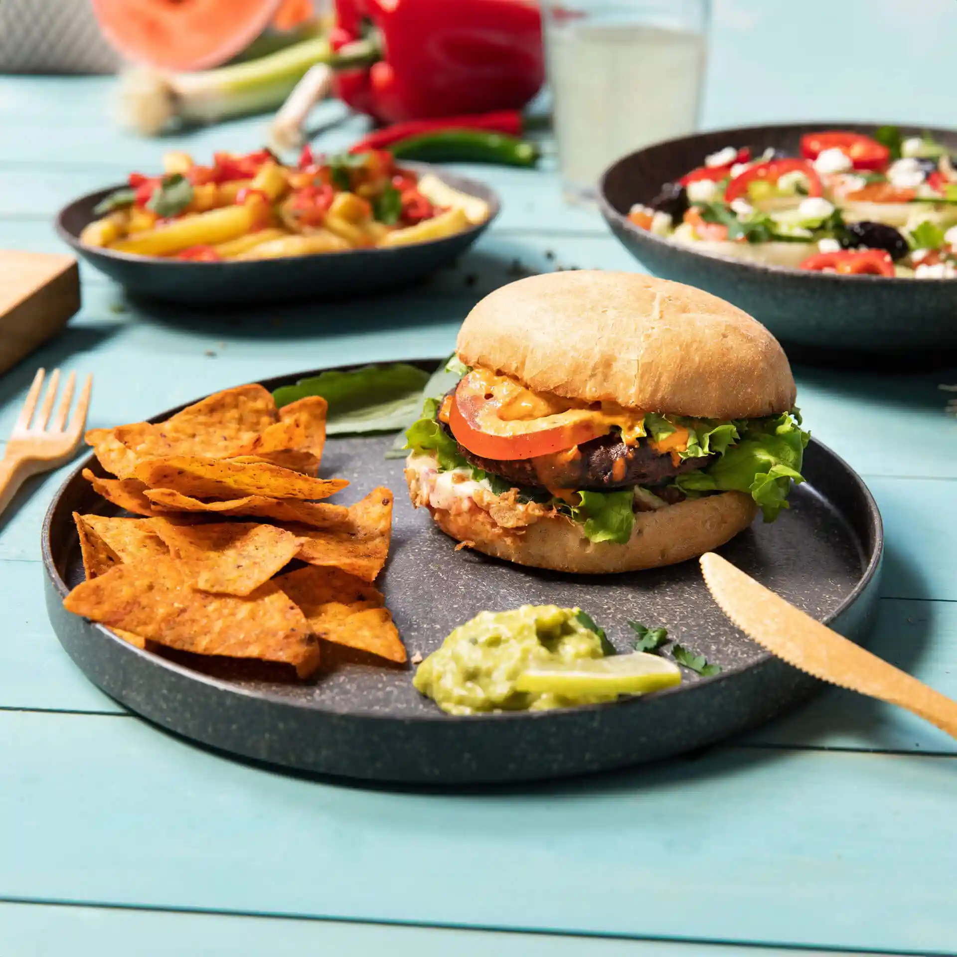 Runde graue wiederverwendbare Platte mit einem Burger, Tortilla-Chips und Guacamole, auf einem blauen Tisch.