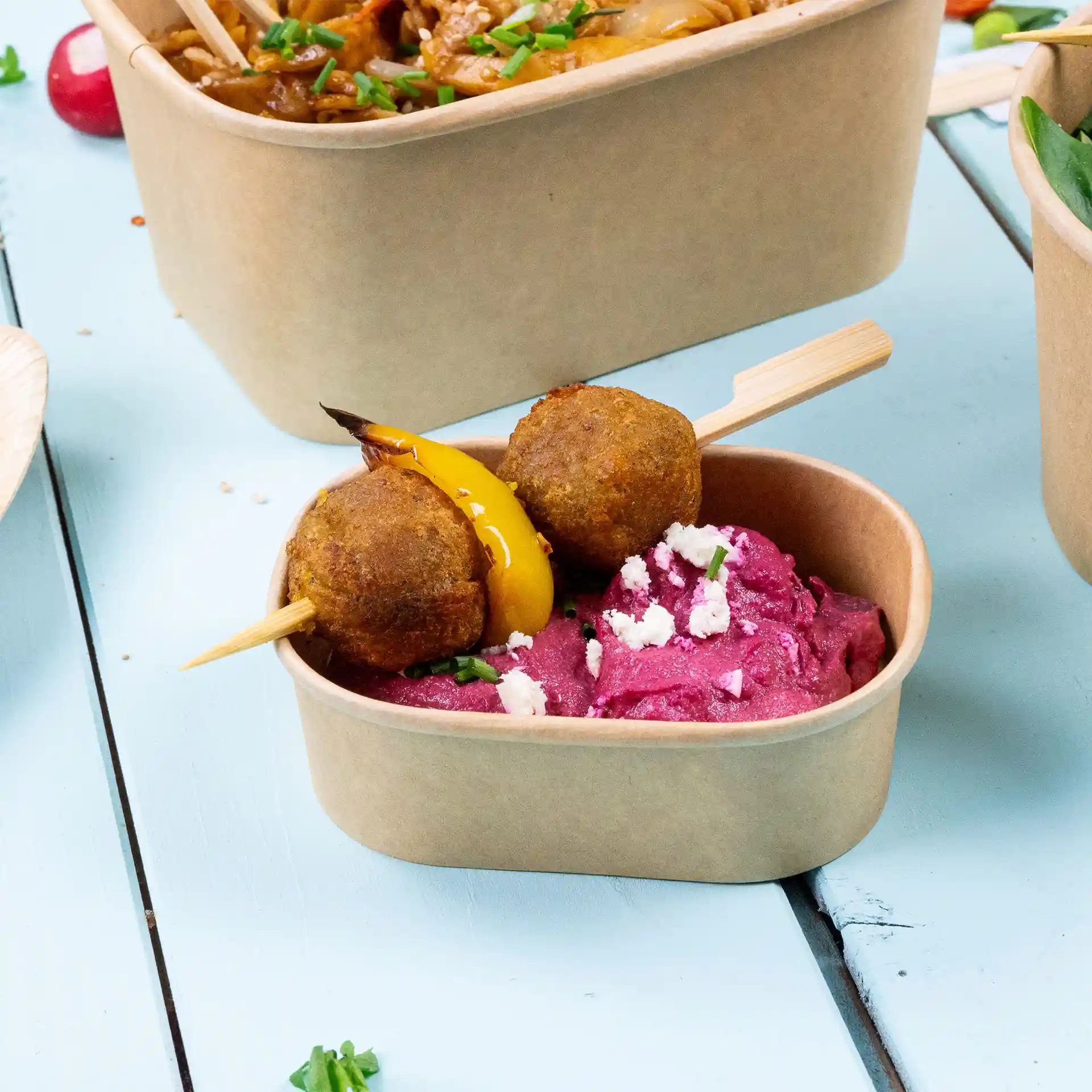 Rectangular kraft food container with pink hummus and two skewered meatballs topped with yellow pepper.