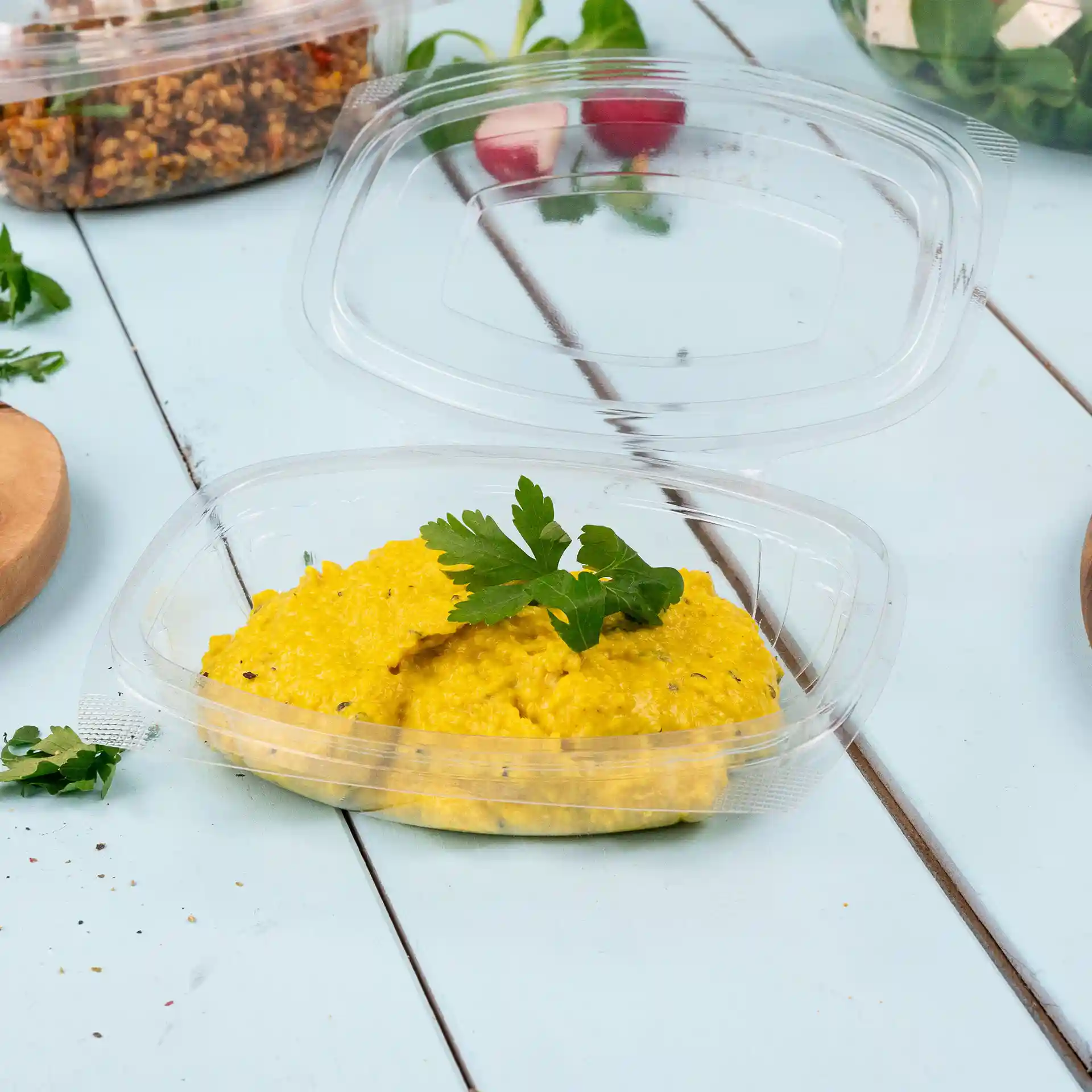 Transparente rechteckige Delikatessenschale mit einem faltbaren Deckel, gefüllt mit gelbem Essen und einem Zweig Petersilie.