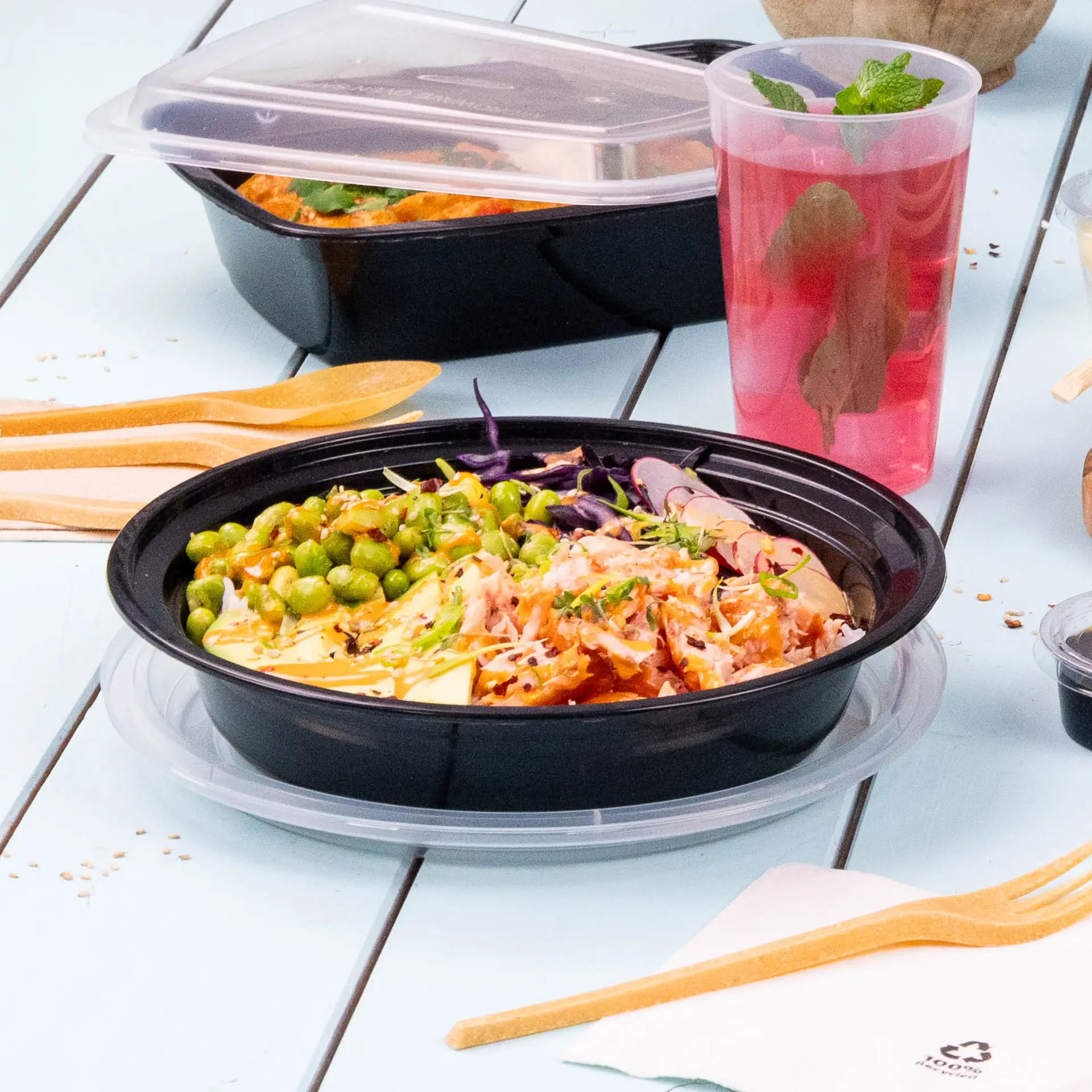 One black reusable plate with mixed food, including peas and shredded vegetables, on a light blue table; wooden utensils nearby.