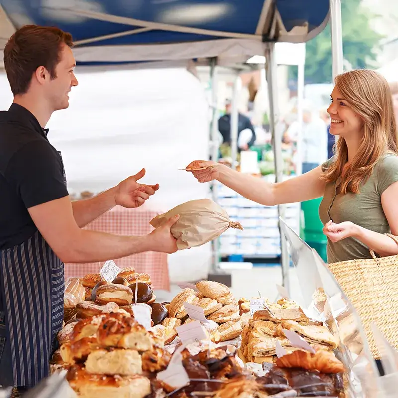 Ein Verkäufer reicht einem Kunden eine braune Papiertüte an einem Marktstand mit verschiedenen Backwaren.