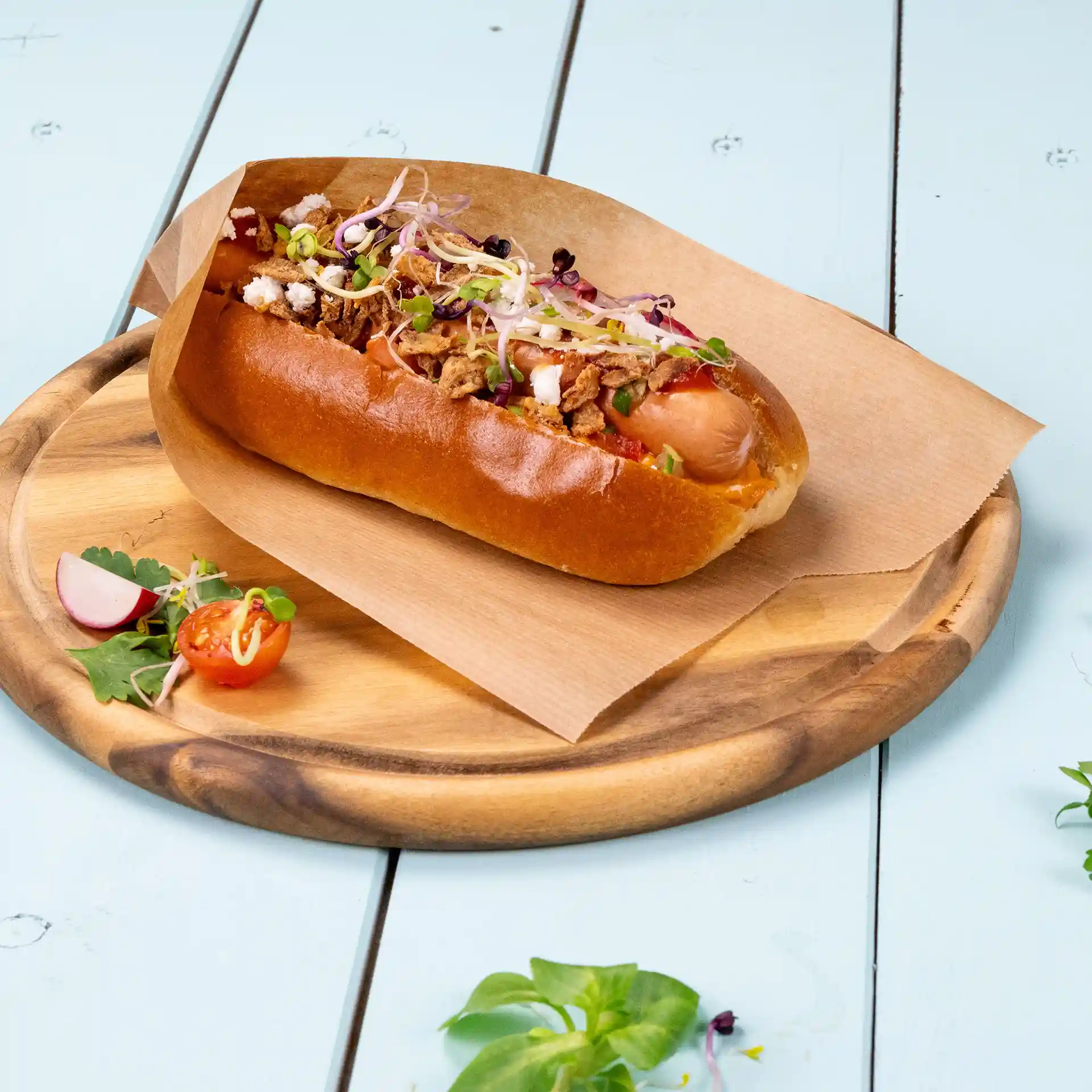 Hot dog in a brown paper wrap on a wooden plate, garnished with vegetables and herbs, with a side of cherry tomatoes and radish.