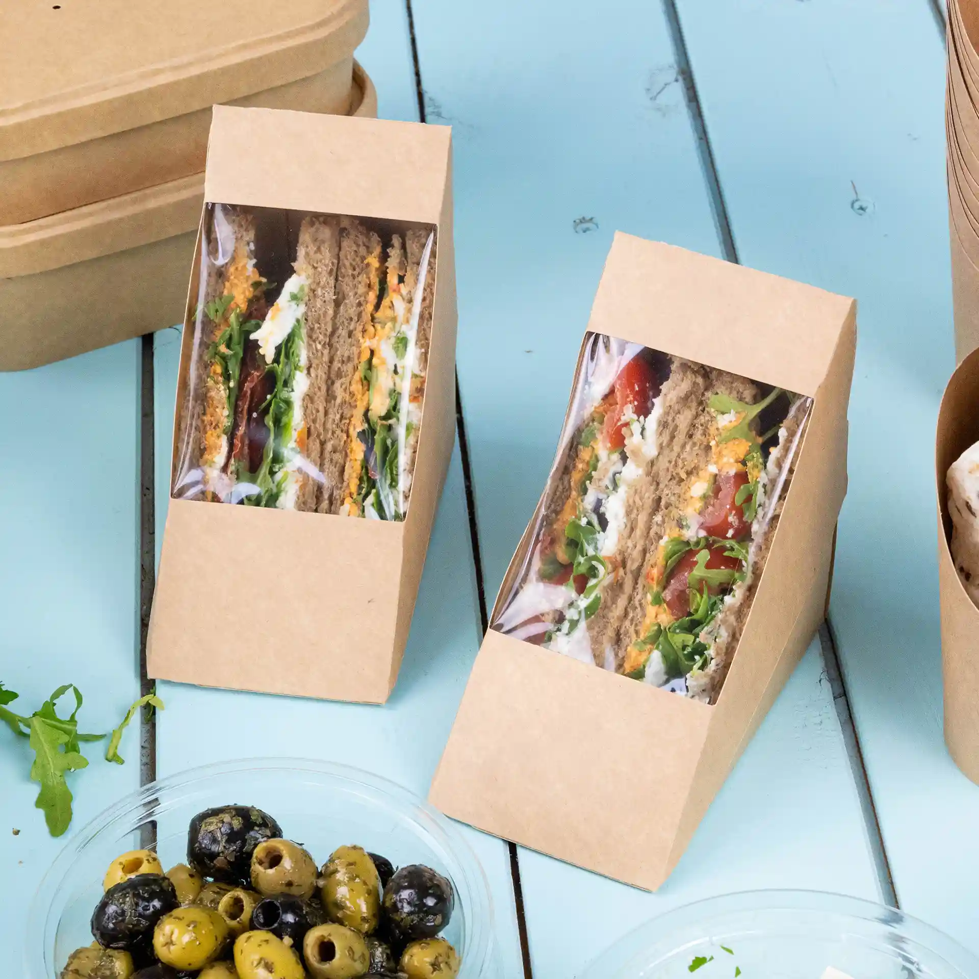 Two triangular cardboard sandwich wedges with a clear viewing window, filled with vegetables, on a blue surface.