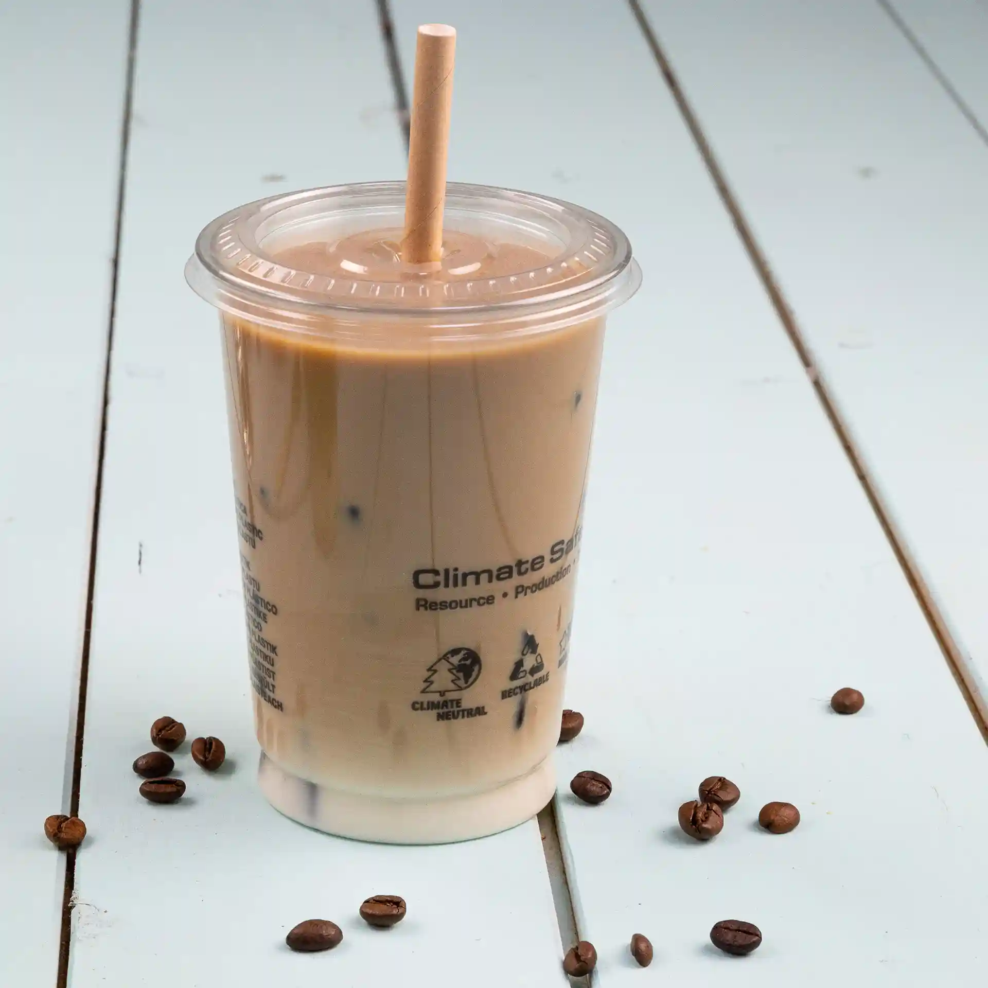 Transparent cup with iced coffee and a paper straw, placed on a light blue surface with scattered coffee beans.