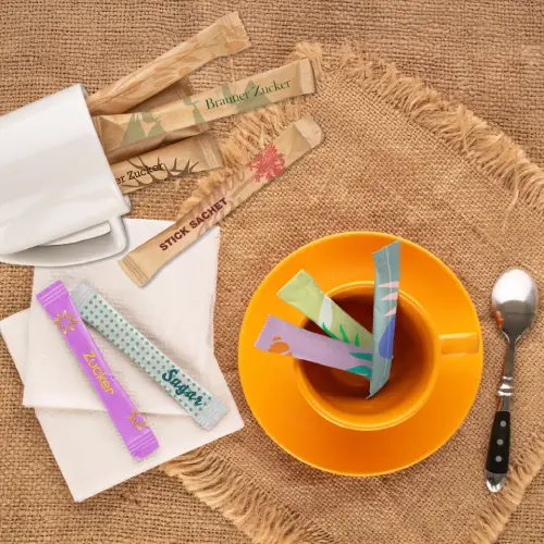 Bunte Zuckersticksäckchen in einer Tasse Tee auf einem rustikalen Tisch mit einem Löffel und leeren Bechern.