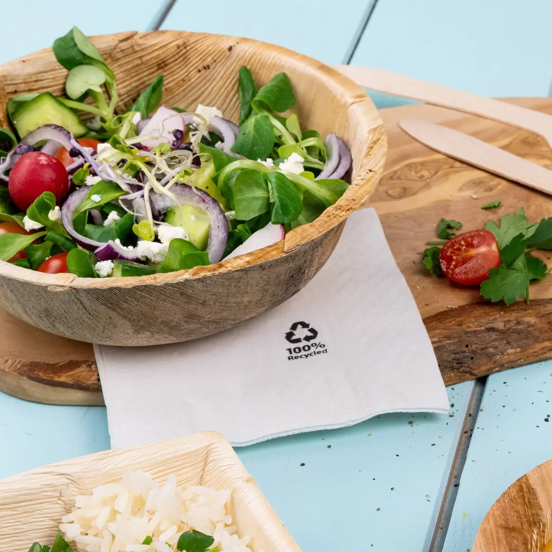 Salad in a wooden bowl with a napkin labeled "100% Recycled," alongside rice and a cherry tomato on a wooden board.