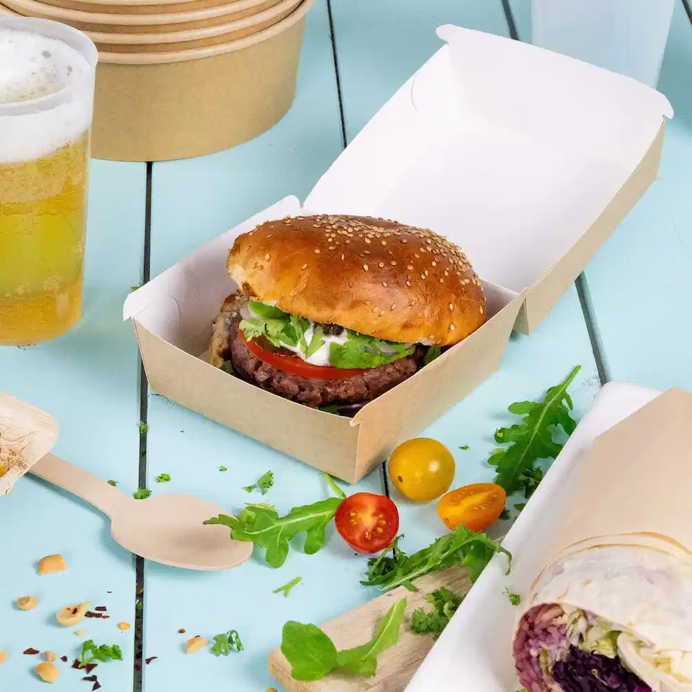Ein Burger mit Sesambrötchen, Salat, Tomate und Sauce in einer braunen Takeout-Box, dazu Gemüse und Bier.