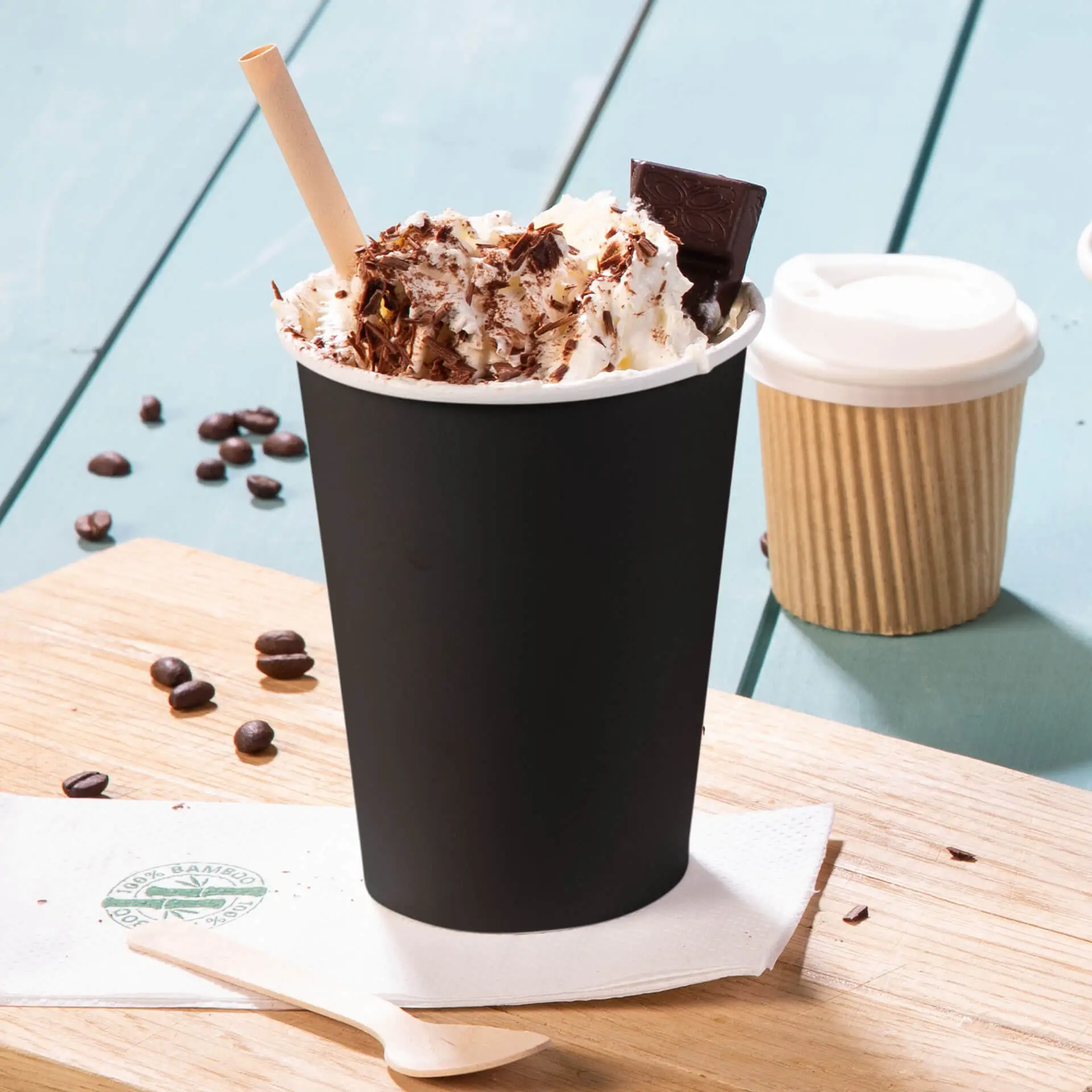 Black single-wall paper cup with whipped cream, chocolate shavings, and a straw, served on a wooden board, with coffee beans nearby.