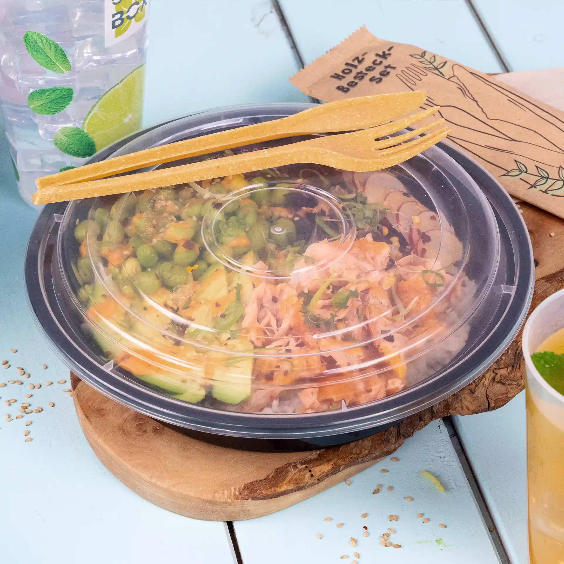 Container with salad, transparent lid, and two brown reusable forks on a wooden board, with a drink and napkin nearby.