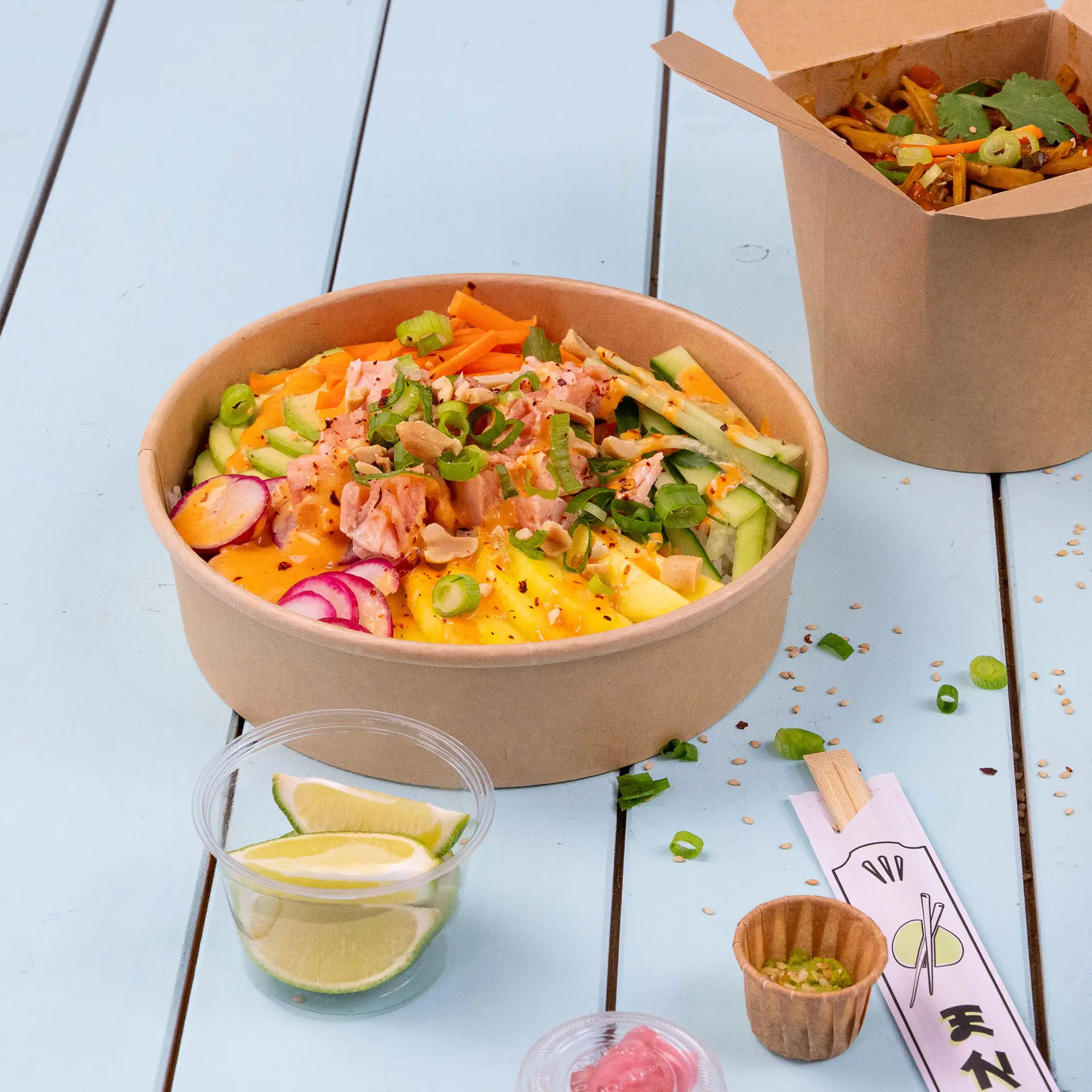 Schüssel gefüllt mit bunten Salatzutaten, neben Limettenscheiben, einem kleinen Behälter mit Wasabi und einer Take-away-Box im Hintergrund.