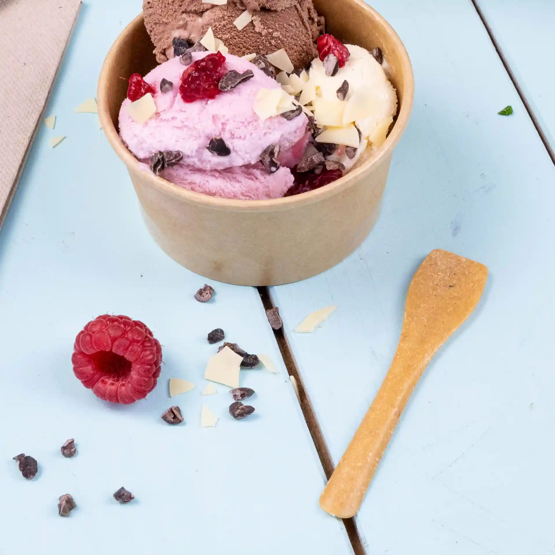 Cup of ice cream with three flavors, raspberries, chocolate chips, and a wooden spoon on a blue surface.