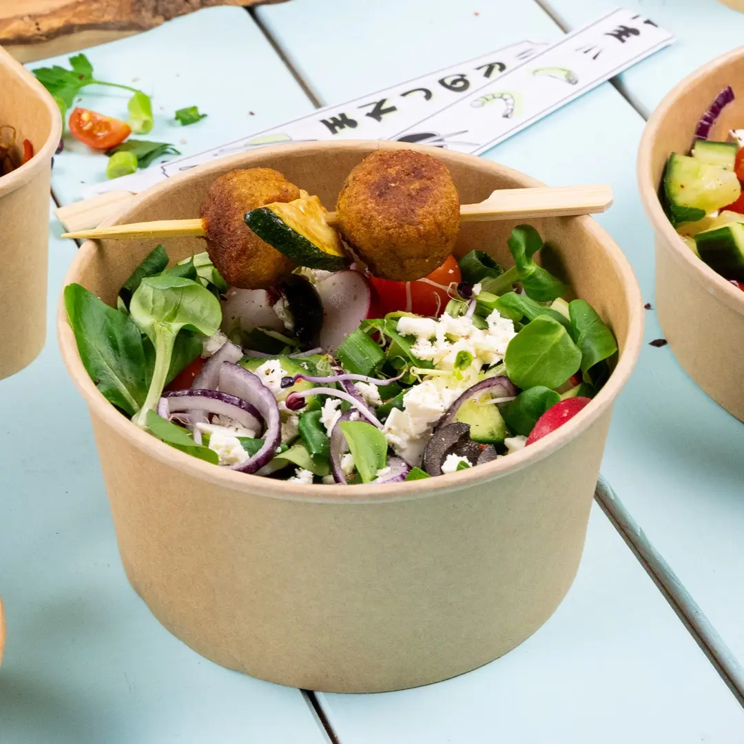 Salad in a brown paper bowl with greens, tomatoes, onions, olives, feta cheese, and two falafel balls on a skewer.