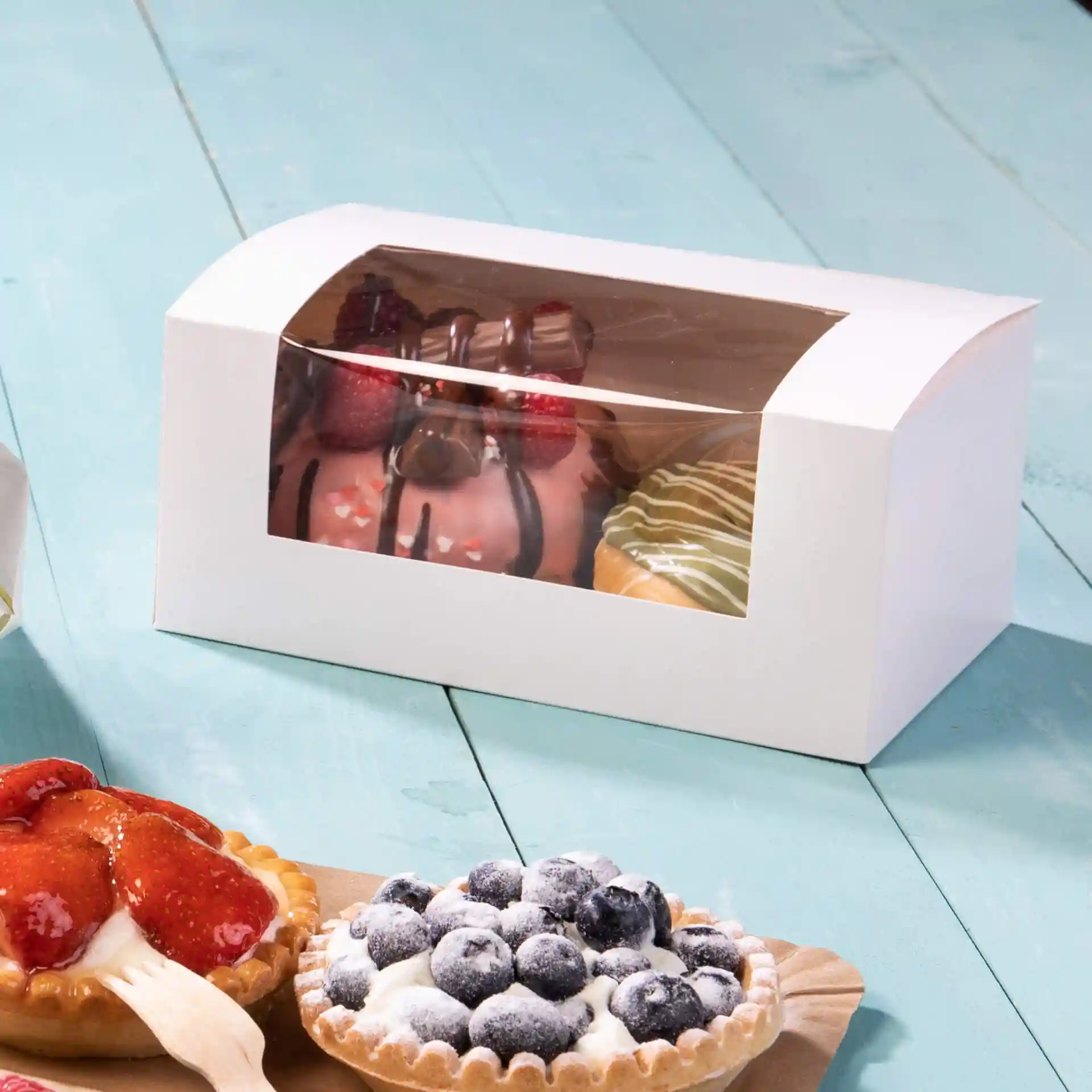 White cardboard cupcake box with a transparent window, displaying decorated cupcakes inside, on a blue surface.