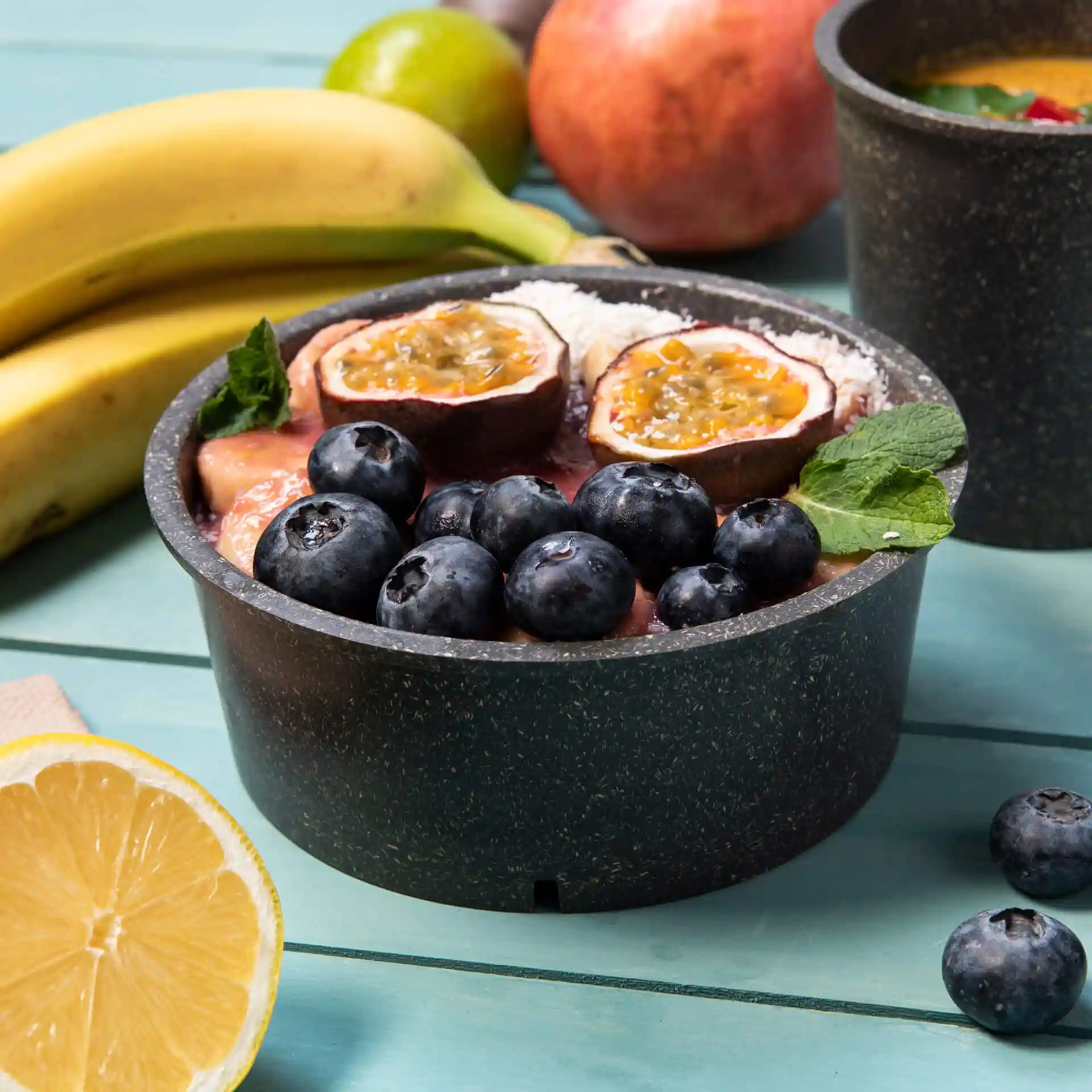 Runde graue wiederverwendbare Schüssel gefüllt mit Obst, darunter Heidelbeeren und Passionsfrucht, auf einer blauen Holzoberfläche.